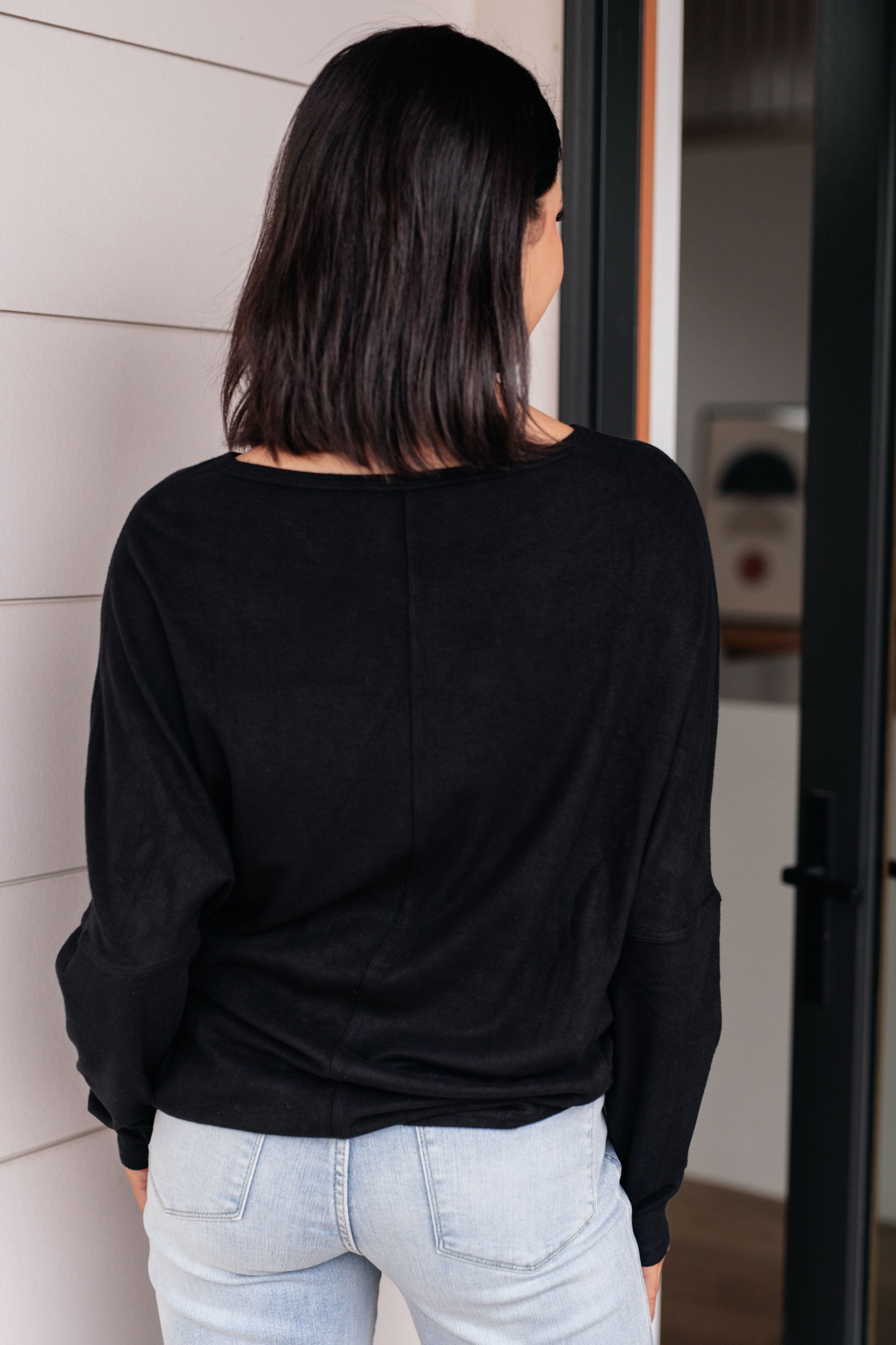 Woman wearing a black dolman long-sleeve top and light blue jeans standing in front of a glass door.