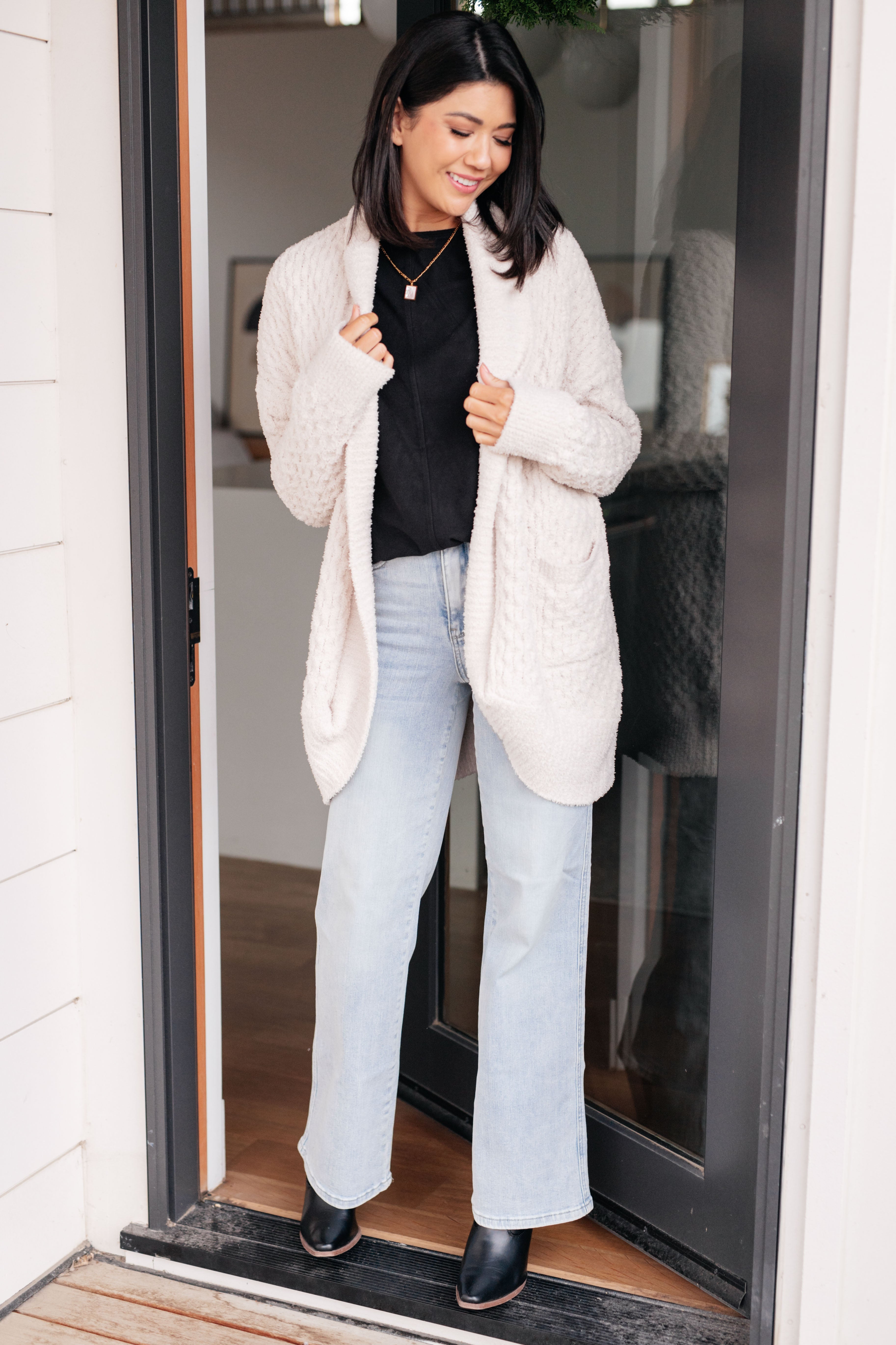 Woman wearing a black dolman long-sleeve top, light blue jeans, black boots and an ivory cardigan standing in front of a glass door.