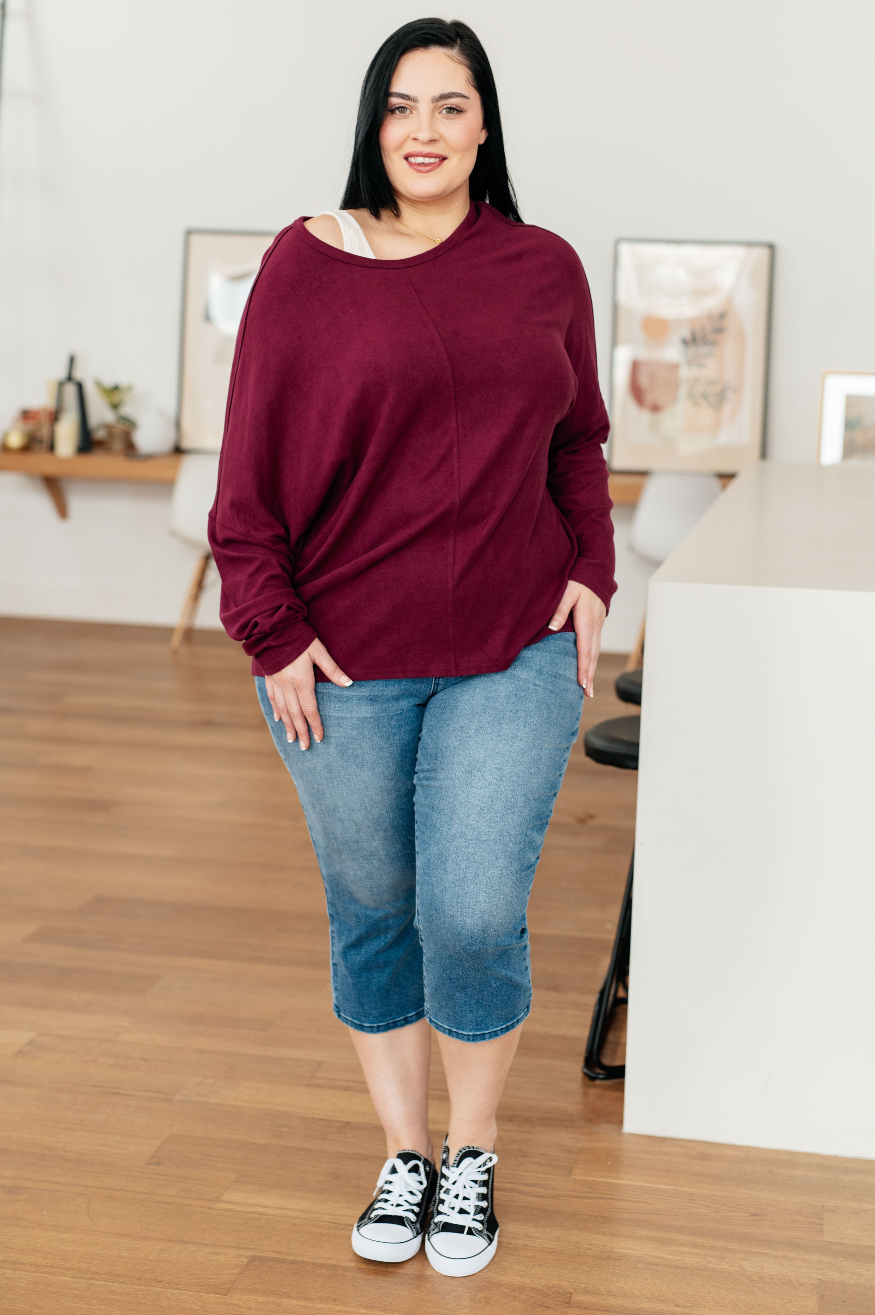 Plus size woman with long black hair wearing wine dolman sleeve top and cropped blue jeans standing indoors