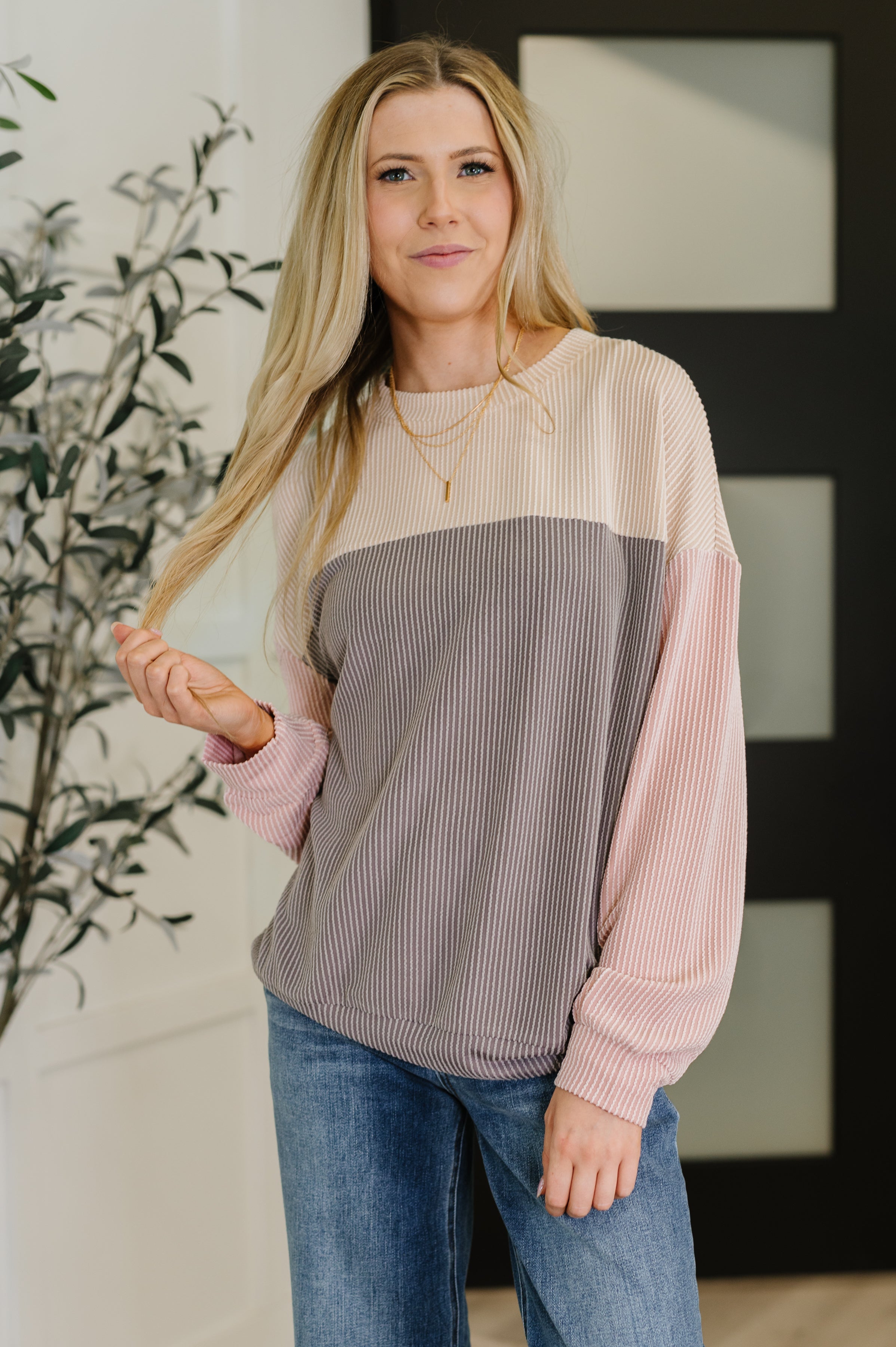 Blonde woman indoors wearing a color-block ribbed knit sweater in cream, gray, and pink with blue jeans