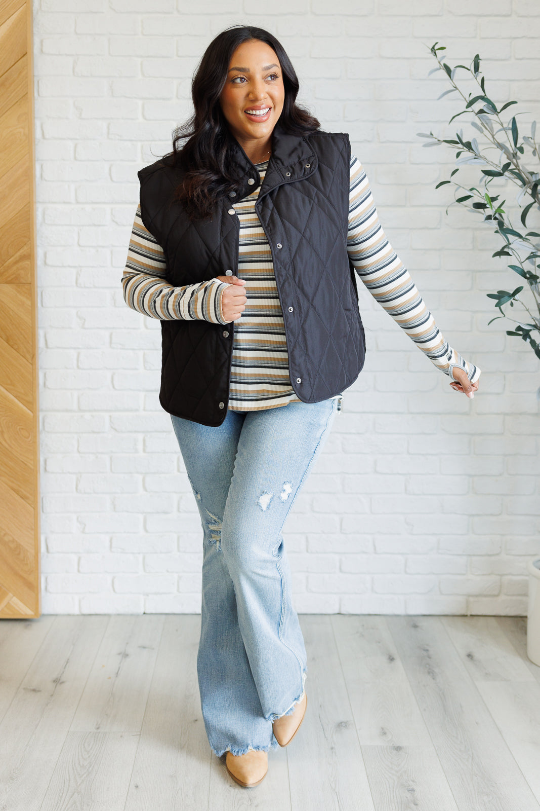 Smiling woman wearing a black quilted vest over a blue striped long-sleeve top and light ripped flared jeans indoors