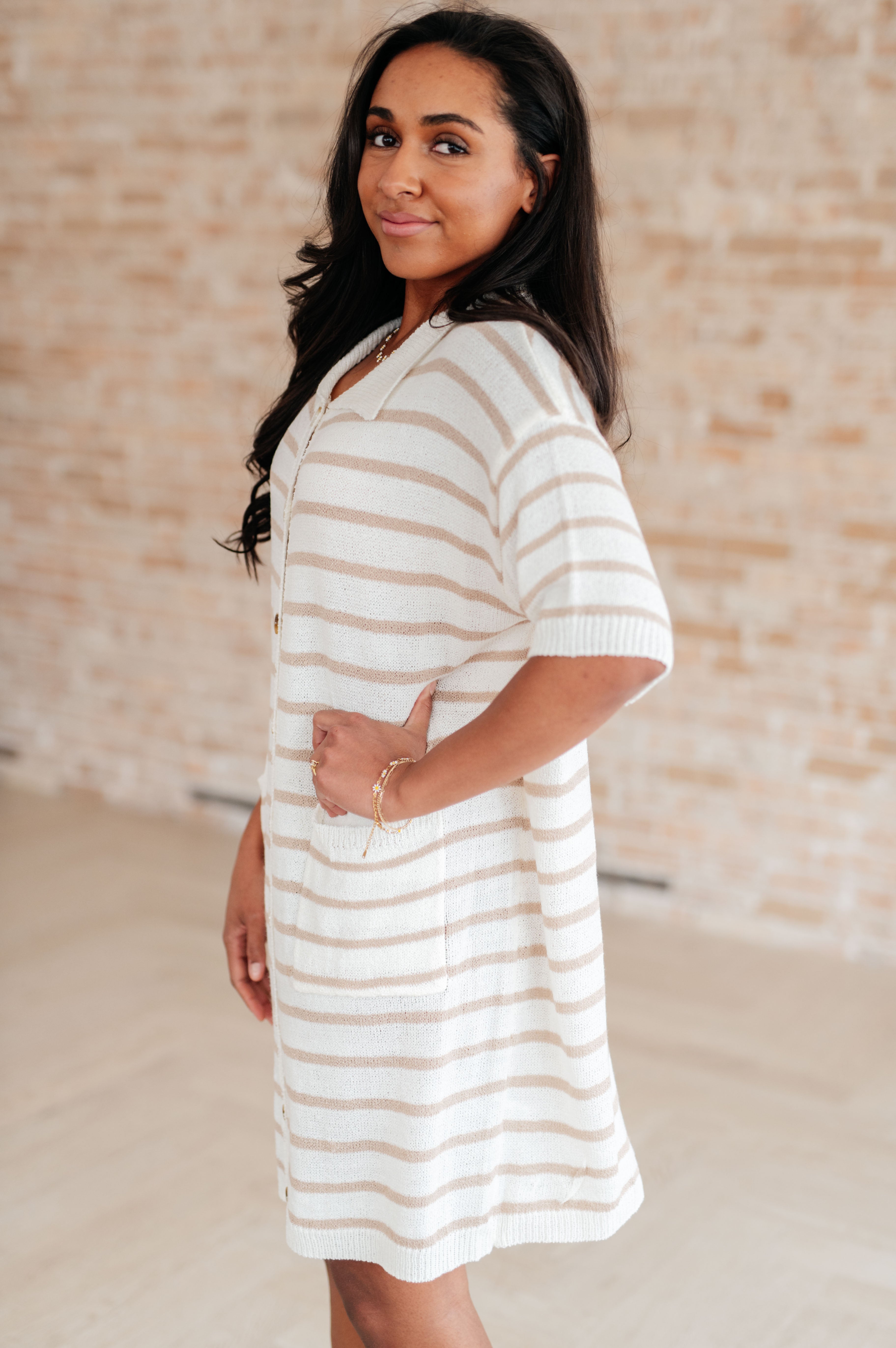Woman wearing beige and white striped button-up collared dress with short sleeves standing sideways indoors