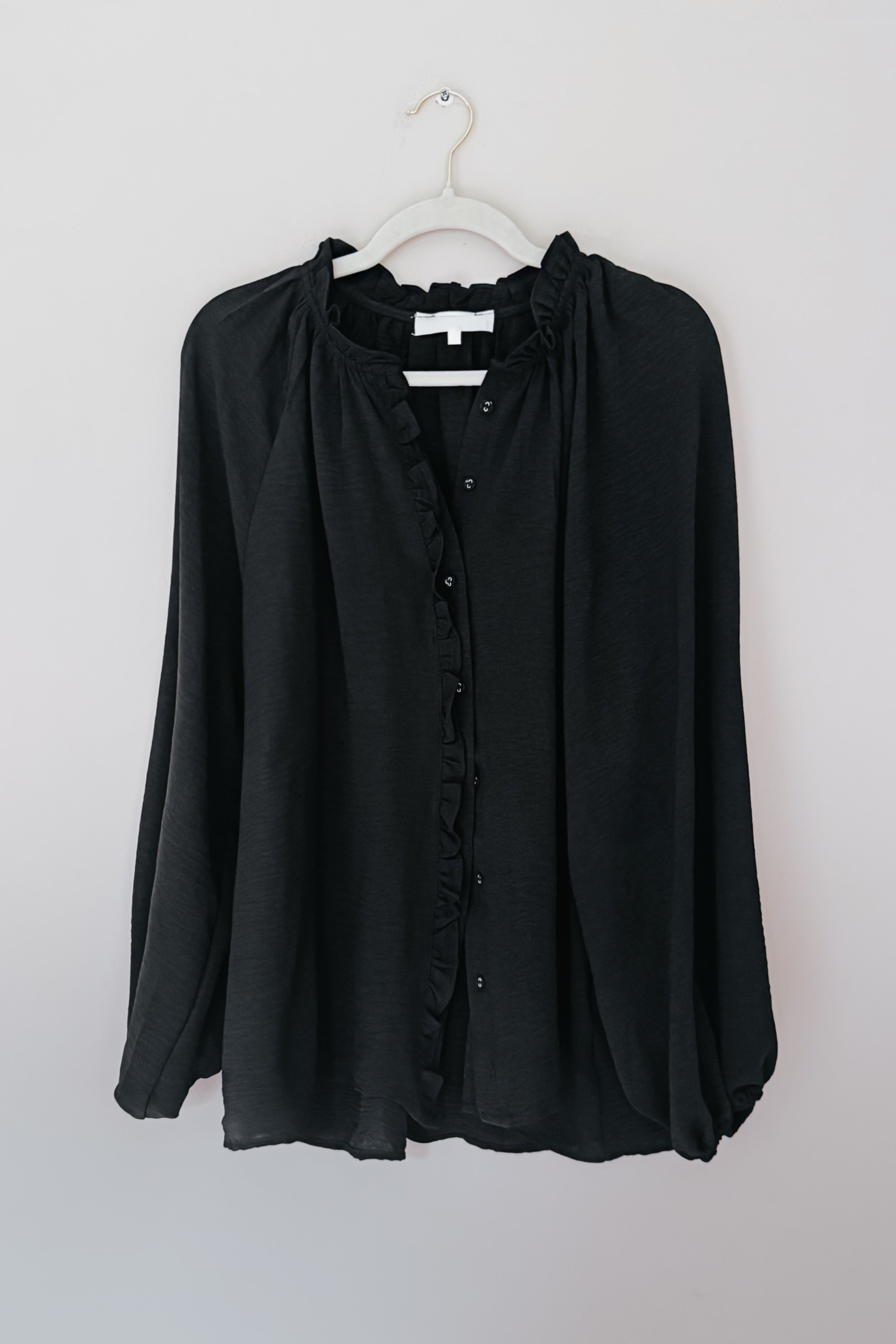 Black button up ruffled blouse on a hanger against a white background