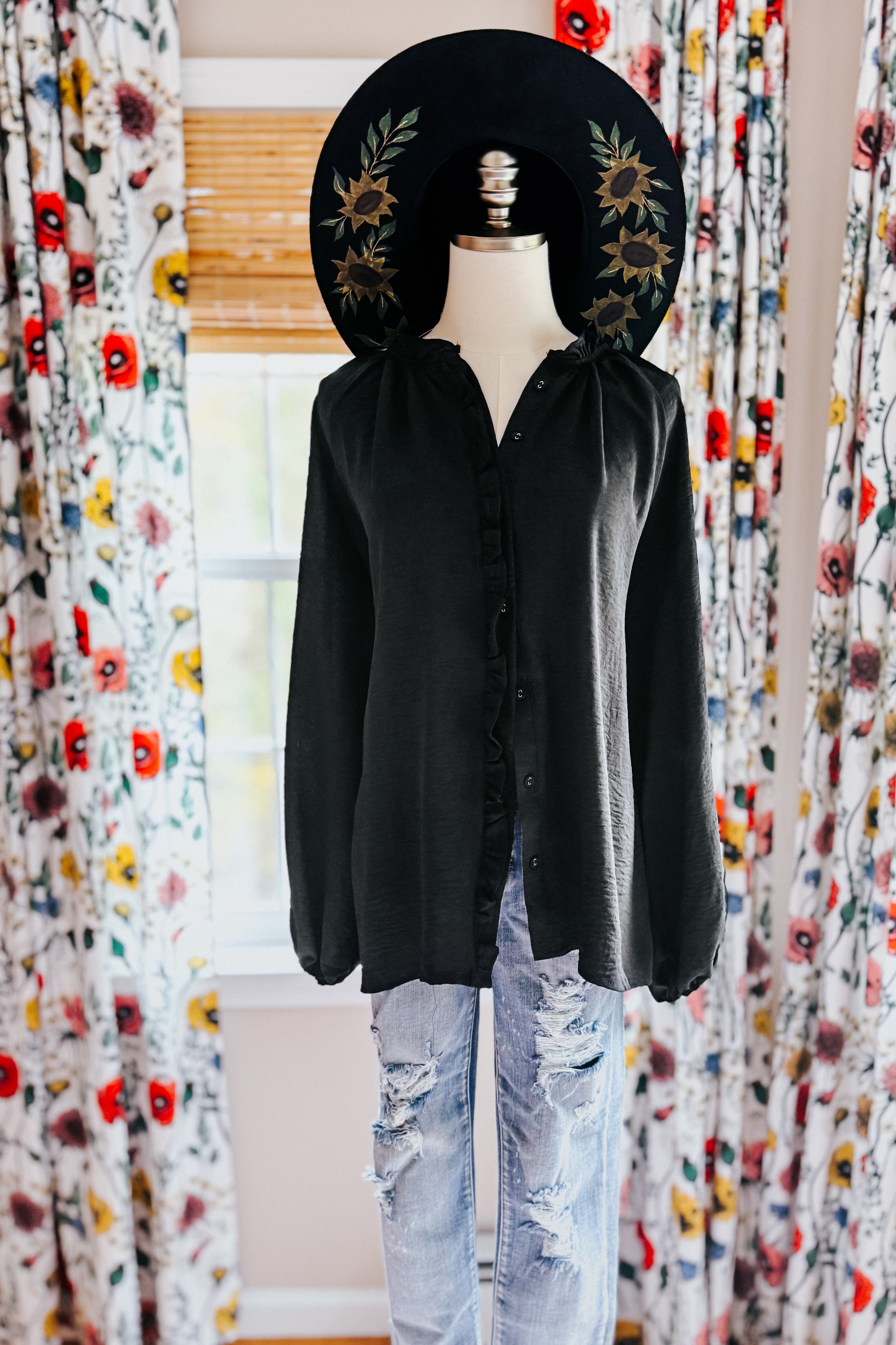 Mannequin wearing a black button up ruffled blouse and jeans with a decorative hat in front of floral curtains.