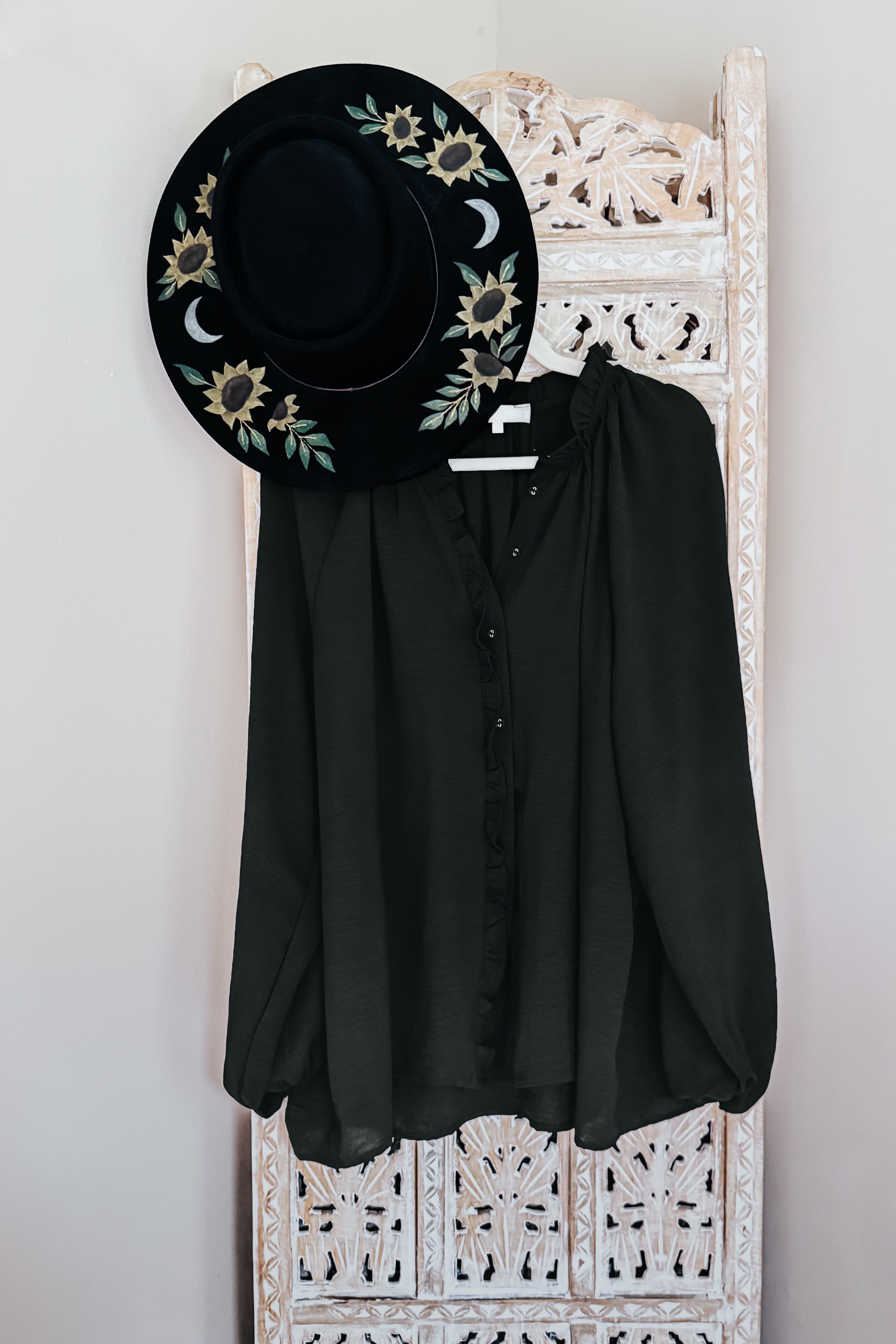 Black hat with floral embroidery and a black button up ruffled blouse on a white decorative rack against a light gray wall.