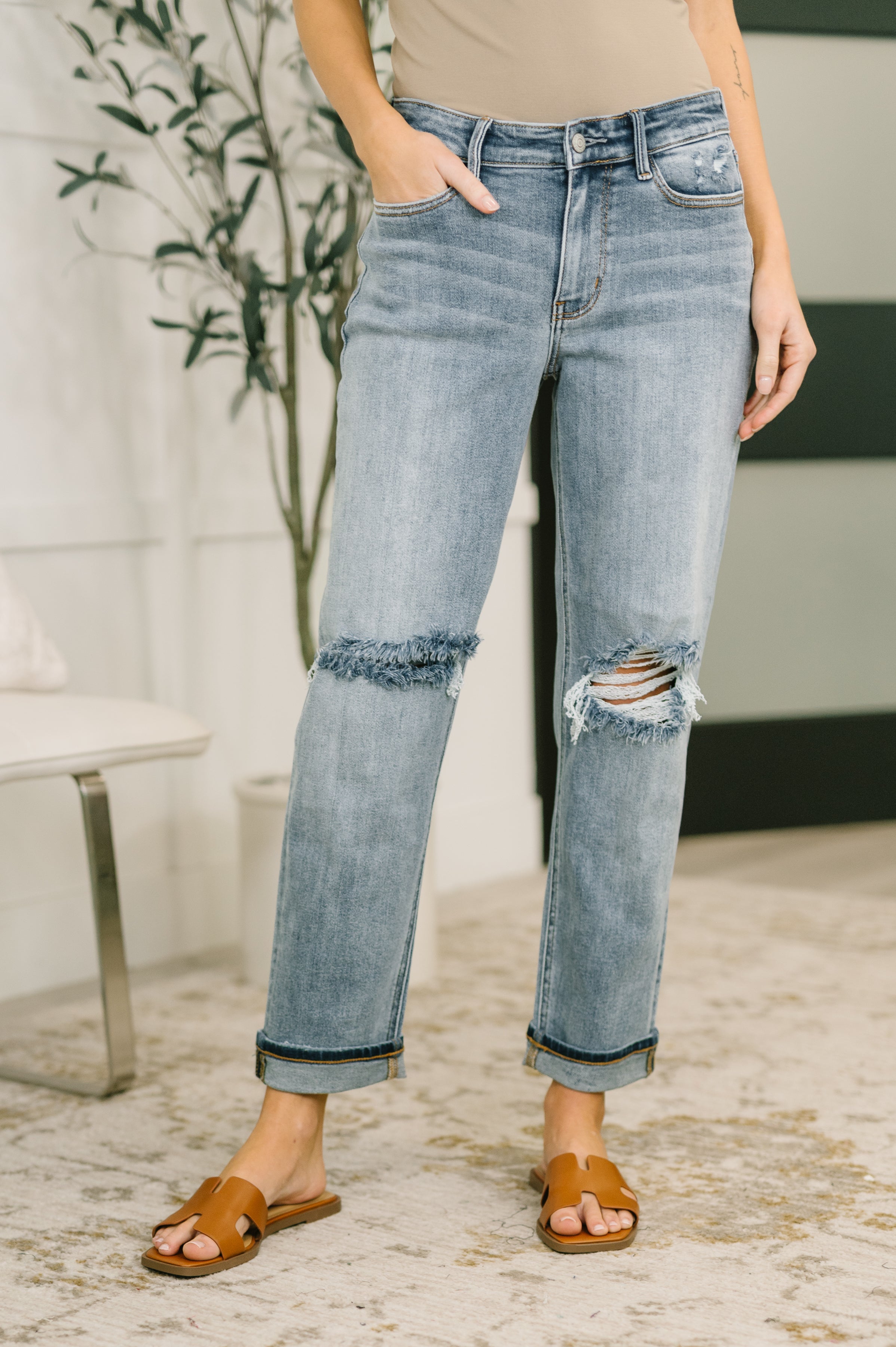 Woman wearing light blue distressed jeans with cuffed hems and brown open-toe flat sandals standing indoors