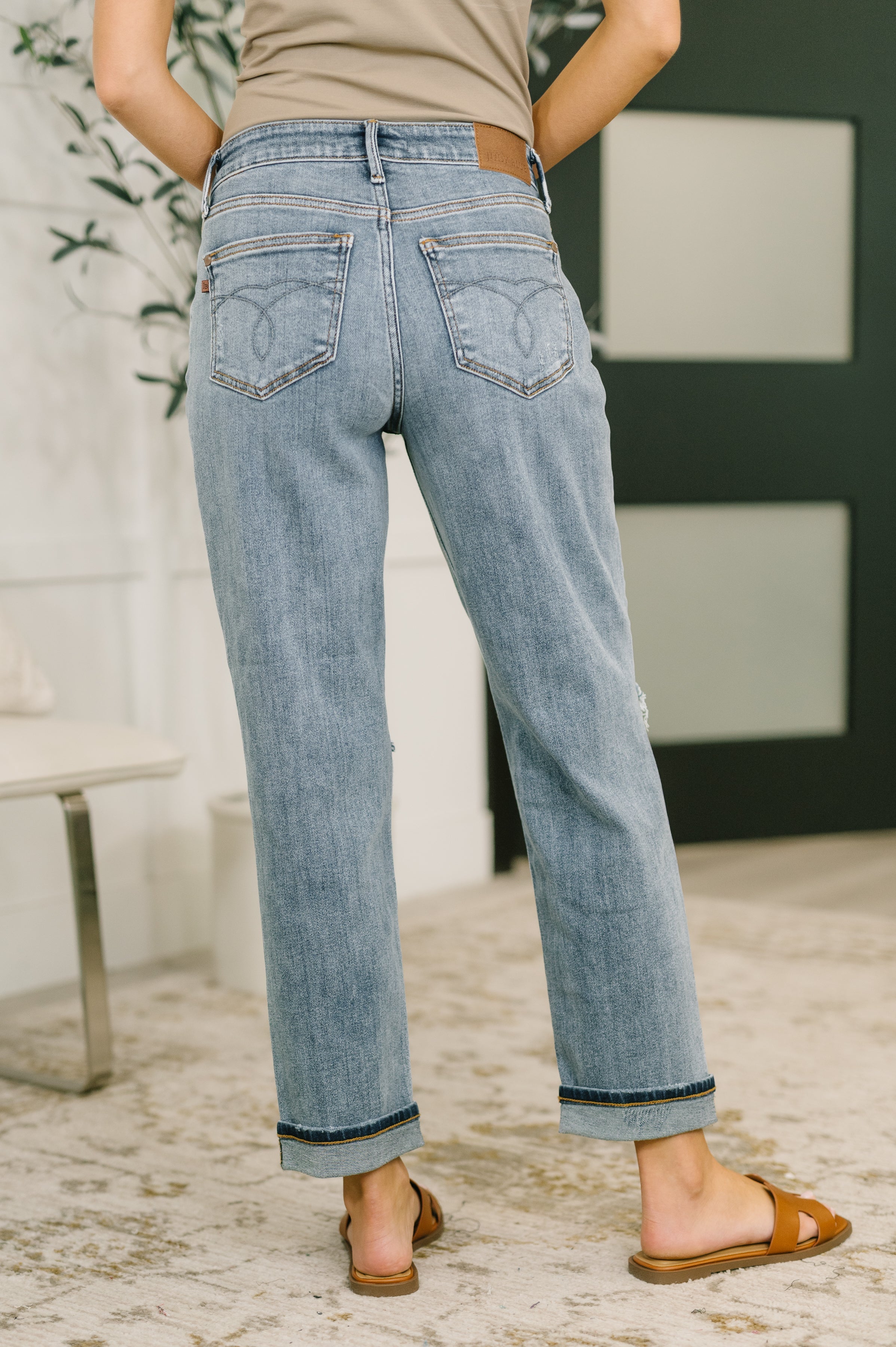 Back view of a person wearing blue straight-leg denim jeans with cuffed hems and brown slip-on sandals indoors