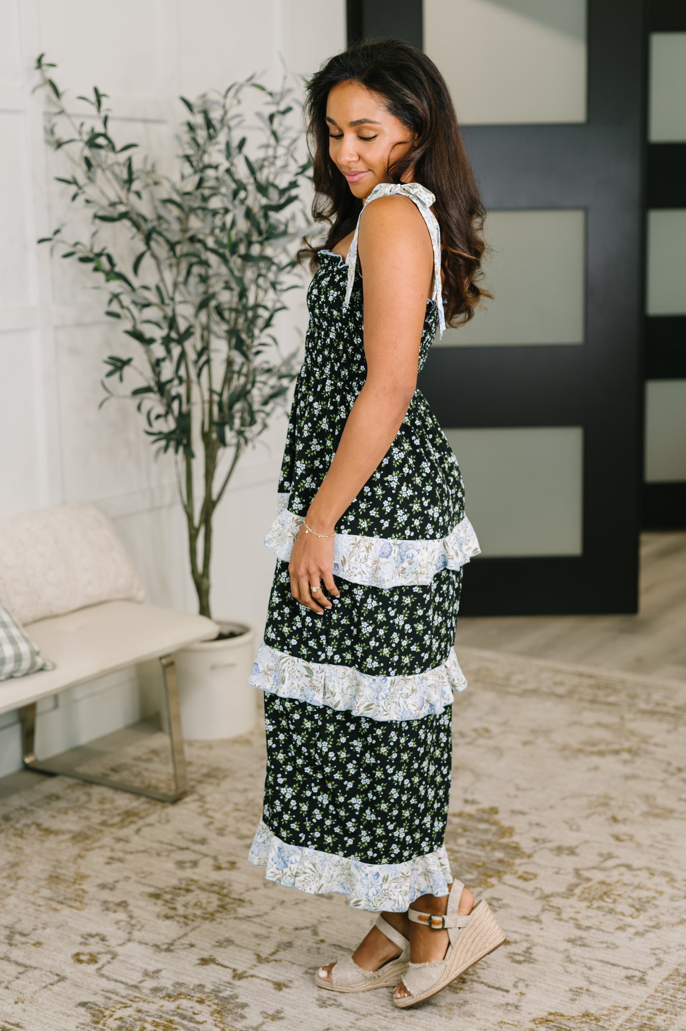 Side view of woman wearing a black floral tiered maxi dress with ruffled light trim and wedge sandals indoors