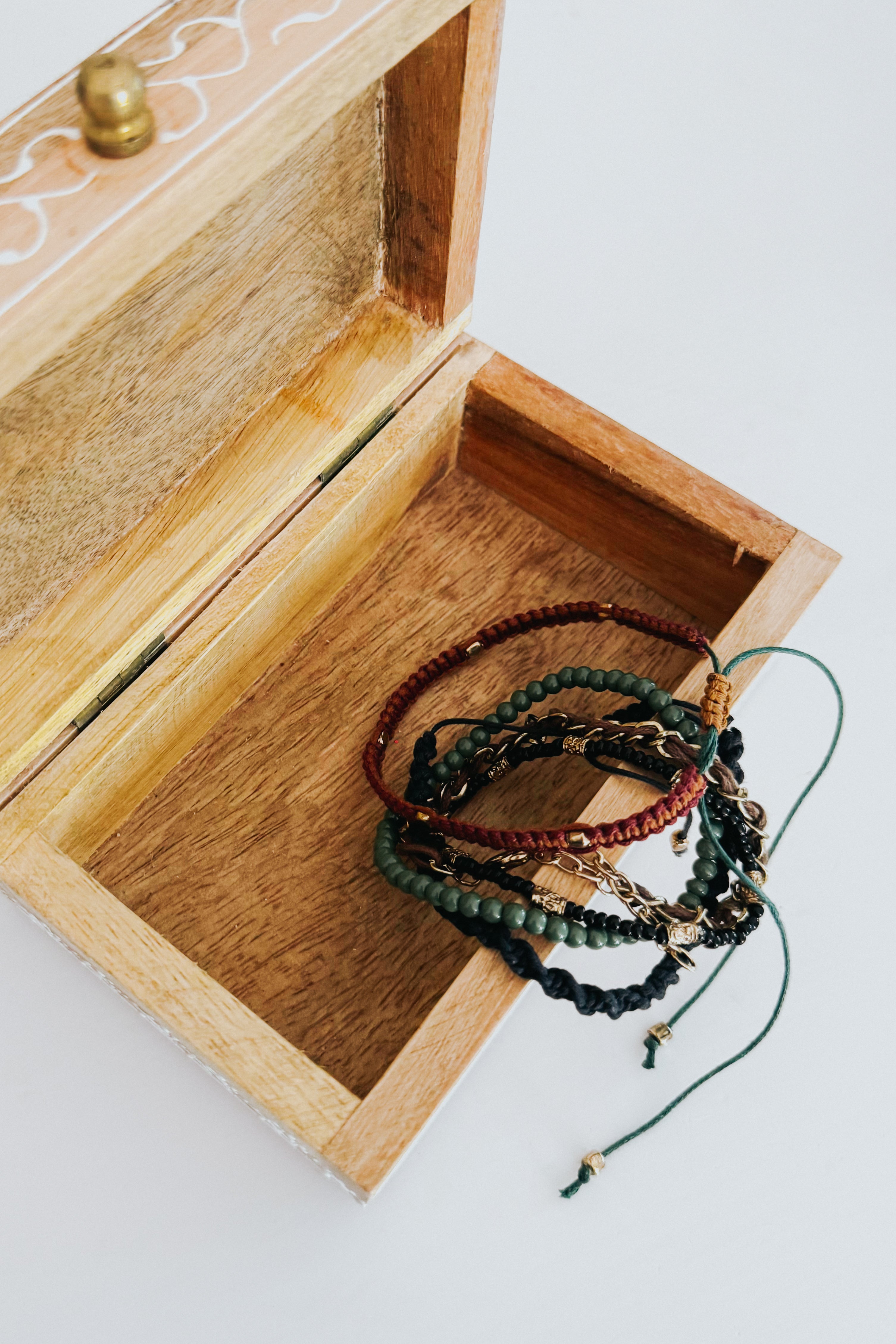Stack of Ember Stackable bracelets with green beads, brown and black braided cords inside an open wooden box on white background