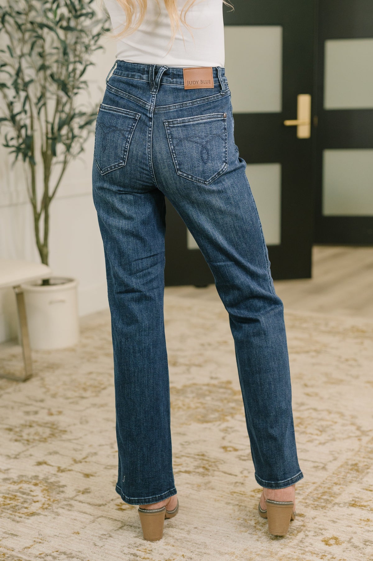 Back view of a woman wearing blue high waist bootcut jeans with welt pockets and tan heeled sandals in a modern entryway