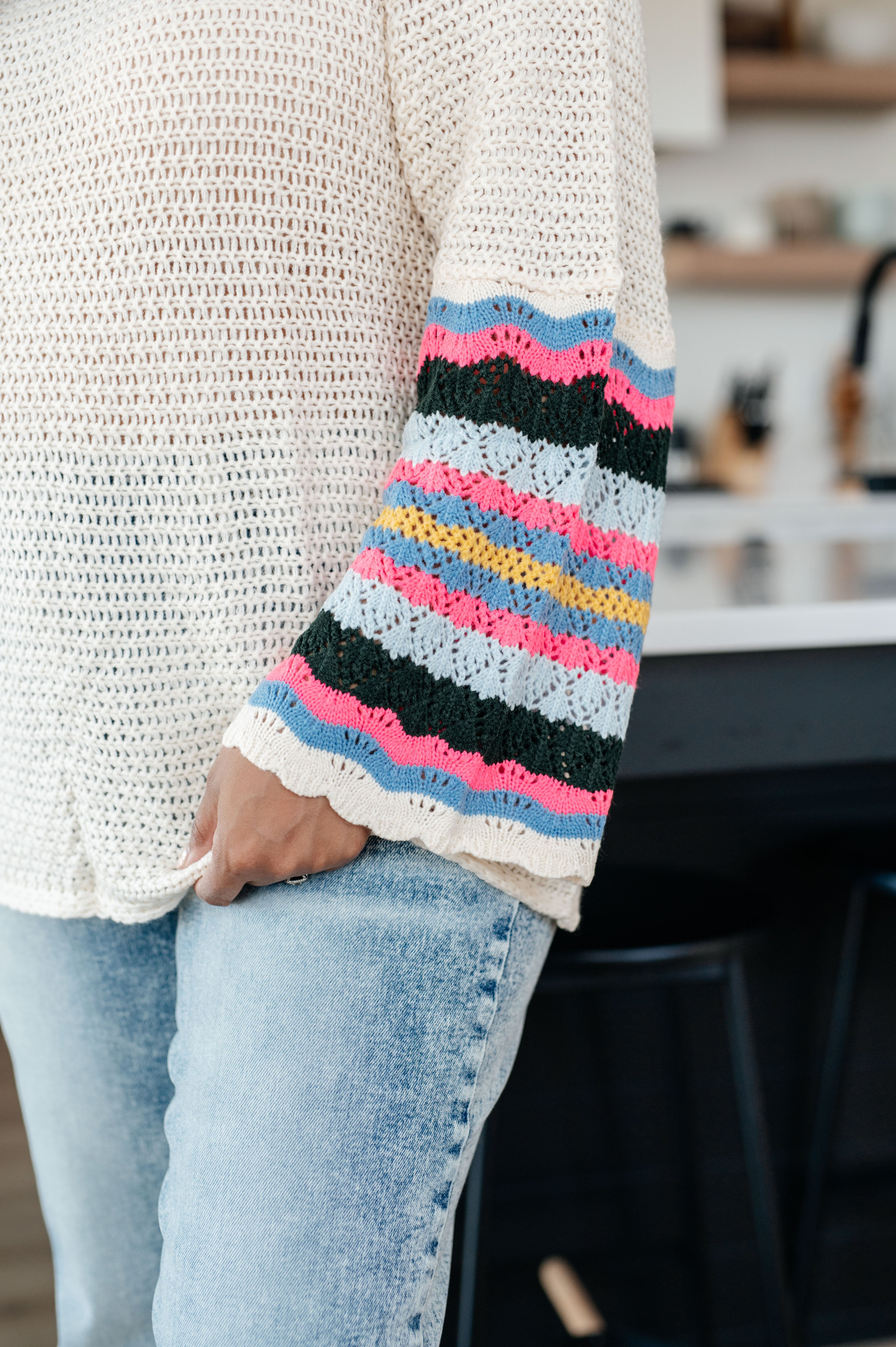 Close-up of cream knit sweater with colorful striped crochet sleeves worn with light blue jeans indoors