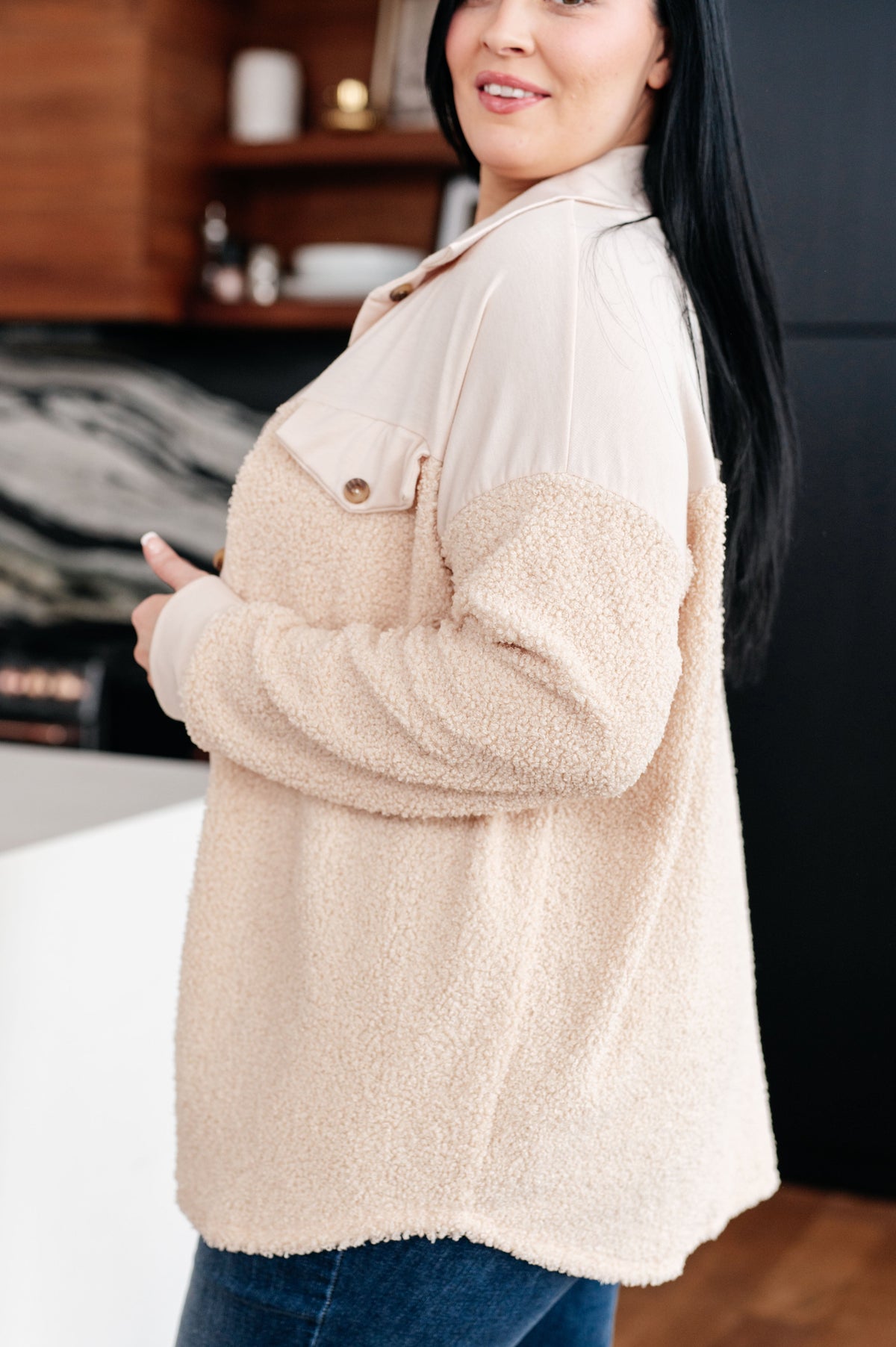 Woman wearing a cream sherpa shacket with a collared neckline and buttoned chest pocket over dark jeans in a modern kitchen