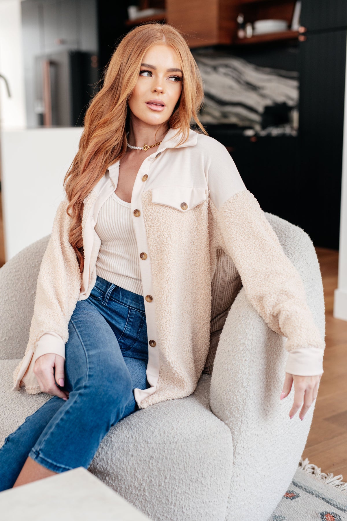 Woman seated indoors wearing a cream sherpa shacket with a collar and faux chest pockets over a ribbed top and blue jeans