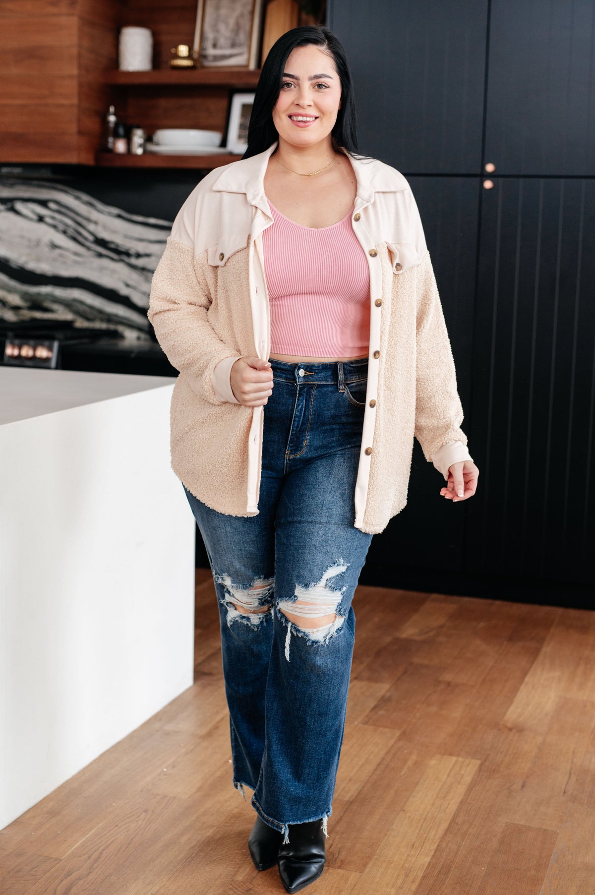 Smiling woman in a cream sherpa shacket over a pink top with distressed jeans and black ankle boots in a modern kitchen
