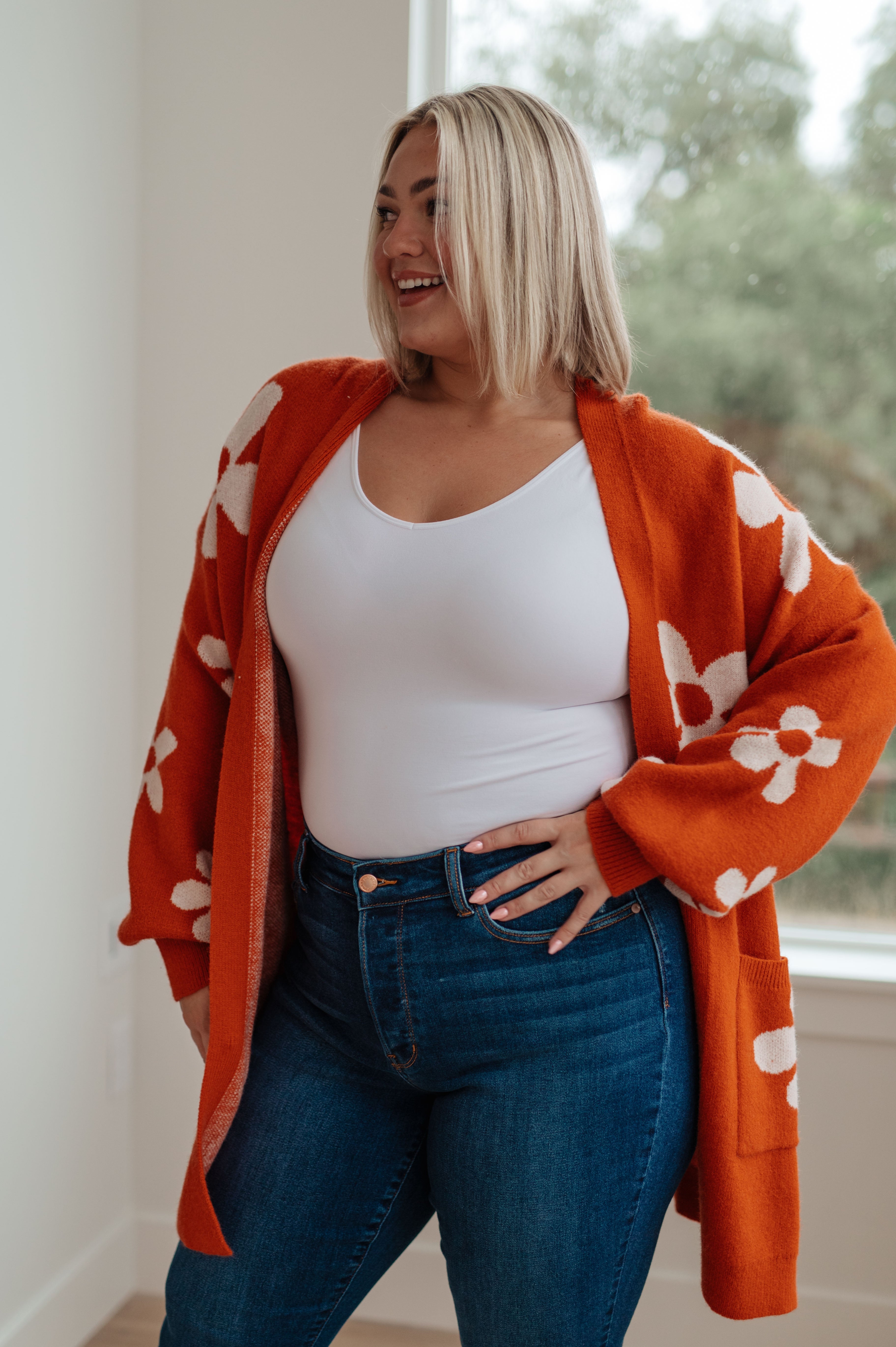 Woman indoors wearing burnt orange floral cardigan over white tank top and high-waisted blue jeans smiling
