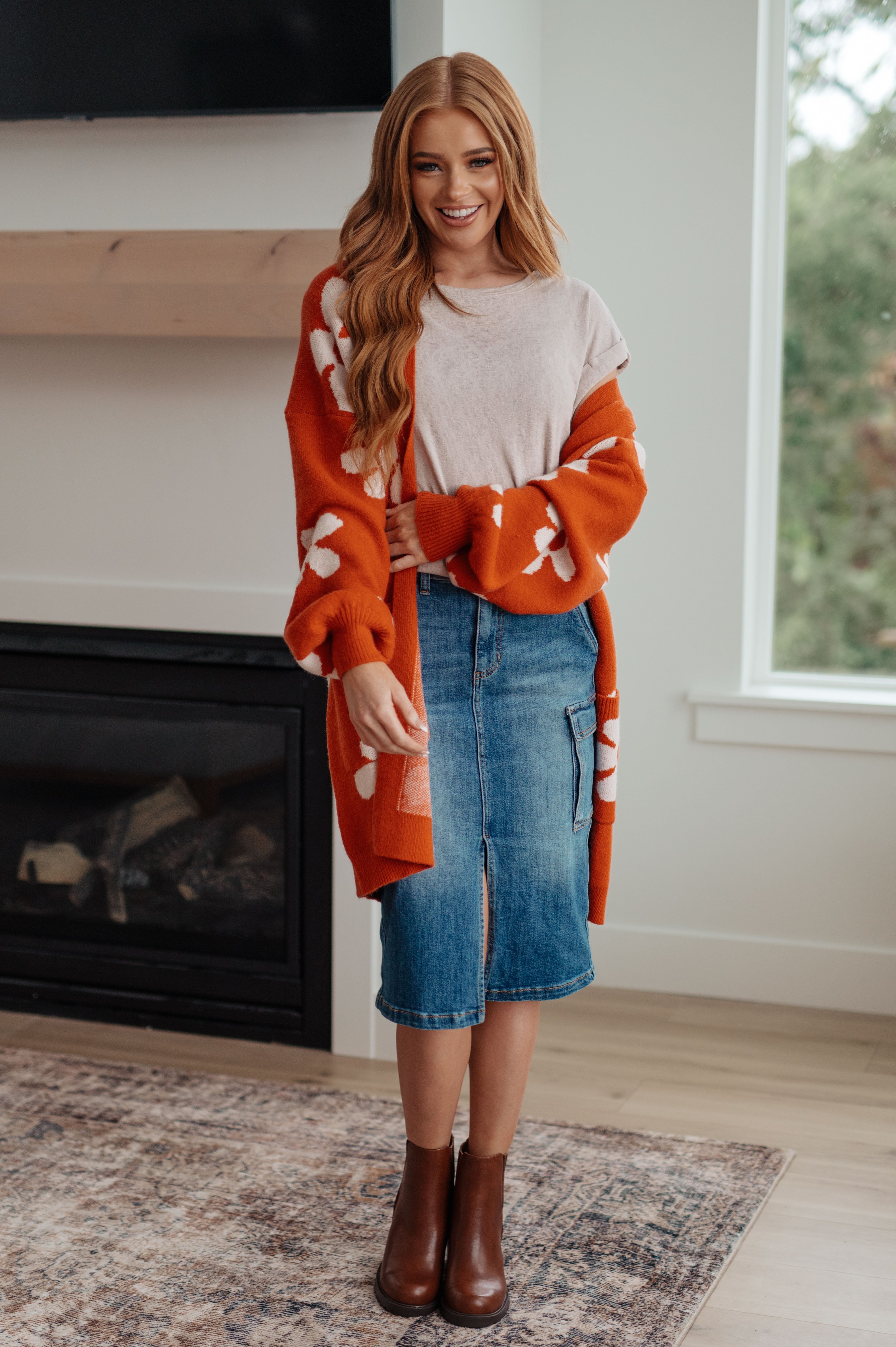 Smiling woman indoors wearing burnt orange floral cardigan over white t-shirt and knee-length denim skirt with brown boots