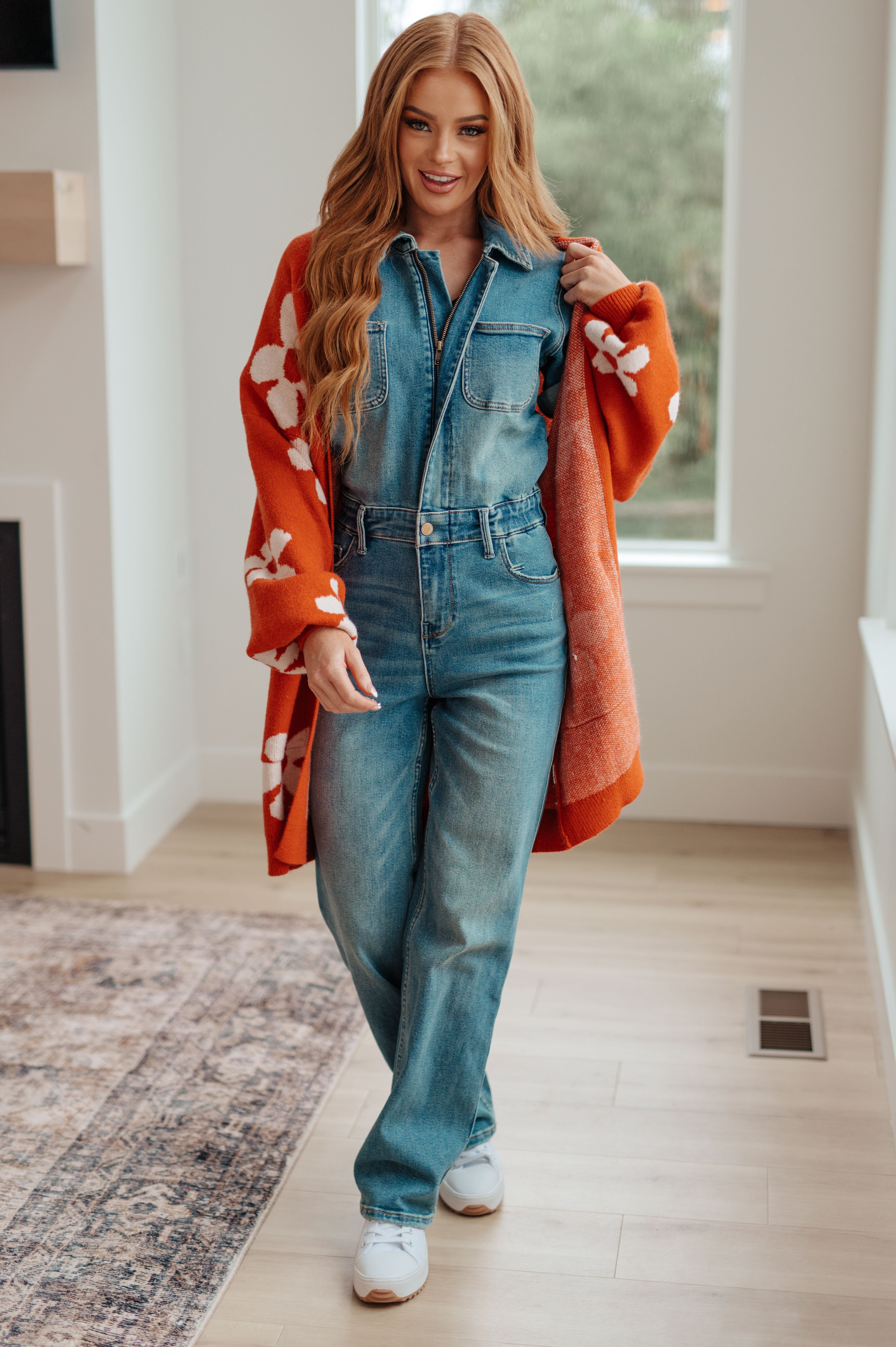 Woman indoors wearing burnt orange floral cardigan over denim jumpsuit and white sneakers walking on wooden floor