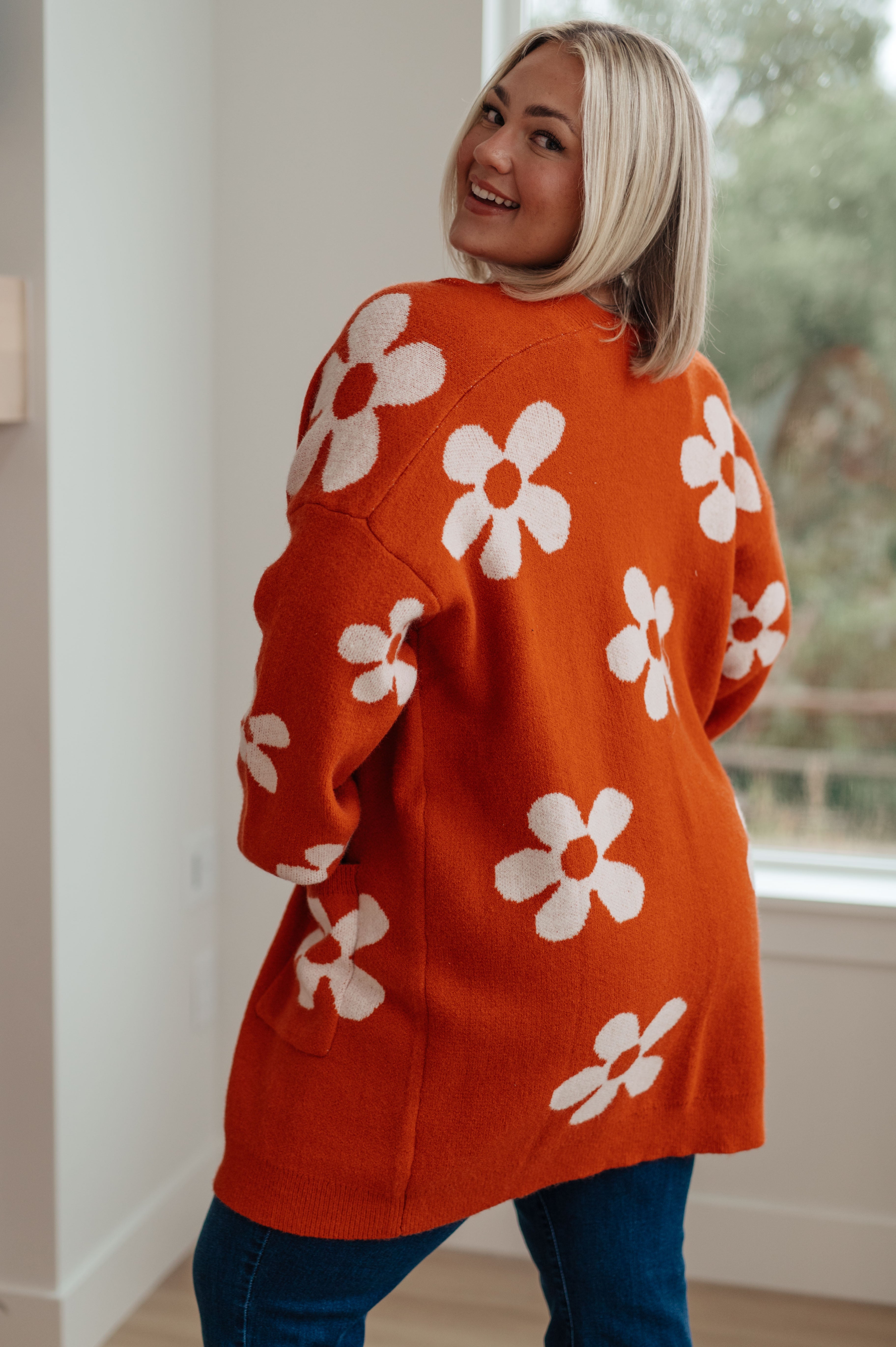 Smiling woman indoors wearing burnt orange cardigan with large white floral patterns paired with dark blue jeans