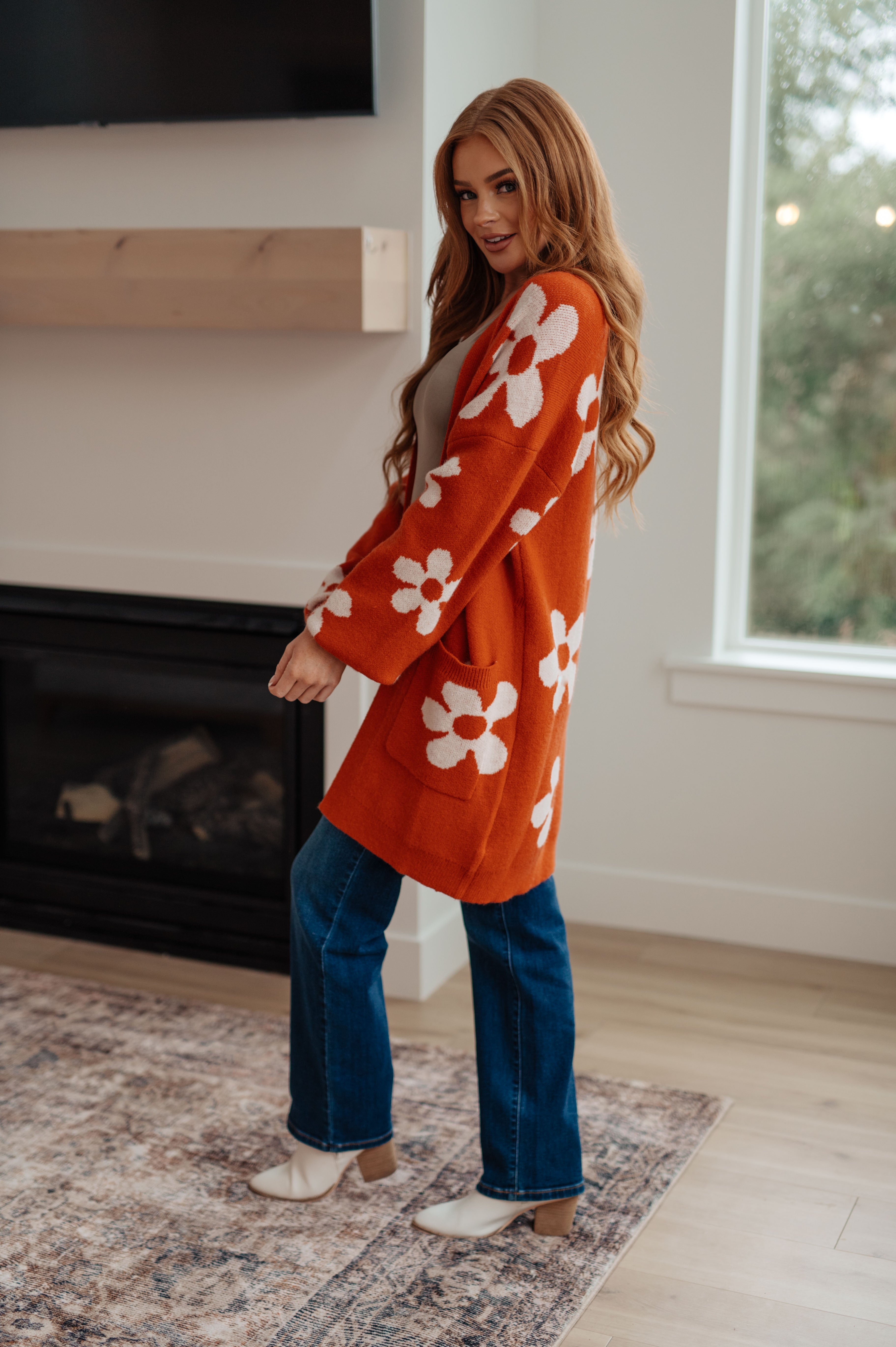Woman indoors wearing burnt orange floral cardigan with pockets and blue jeans standing near fireplace and window
