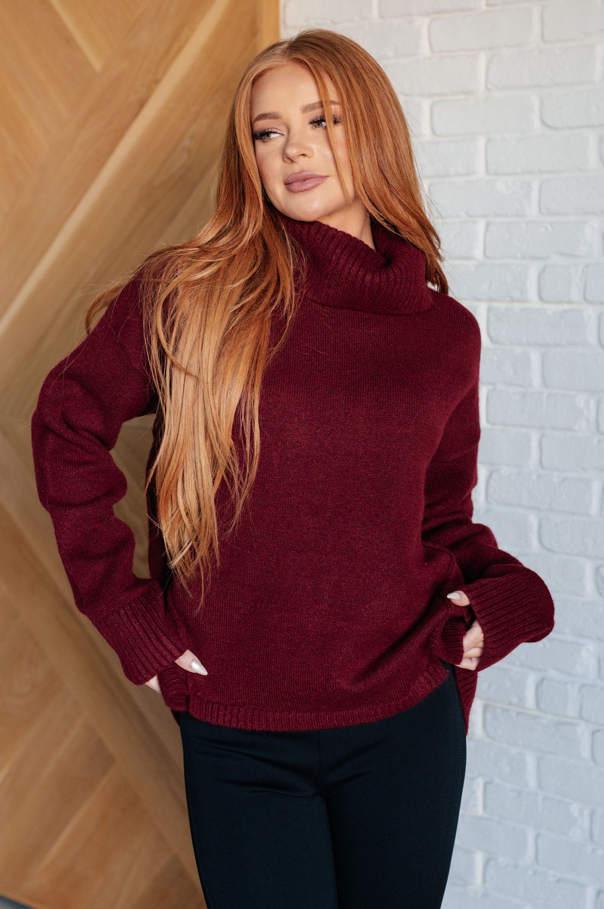 Woman wearing a burgundy turtleneck sweater with drop shoulders, ribbed cuffs, and a split stepped hem with black pants indoors