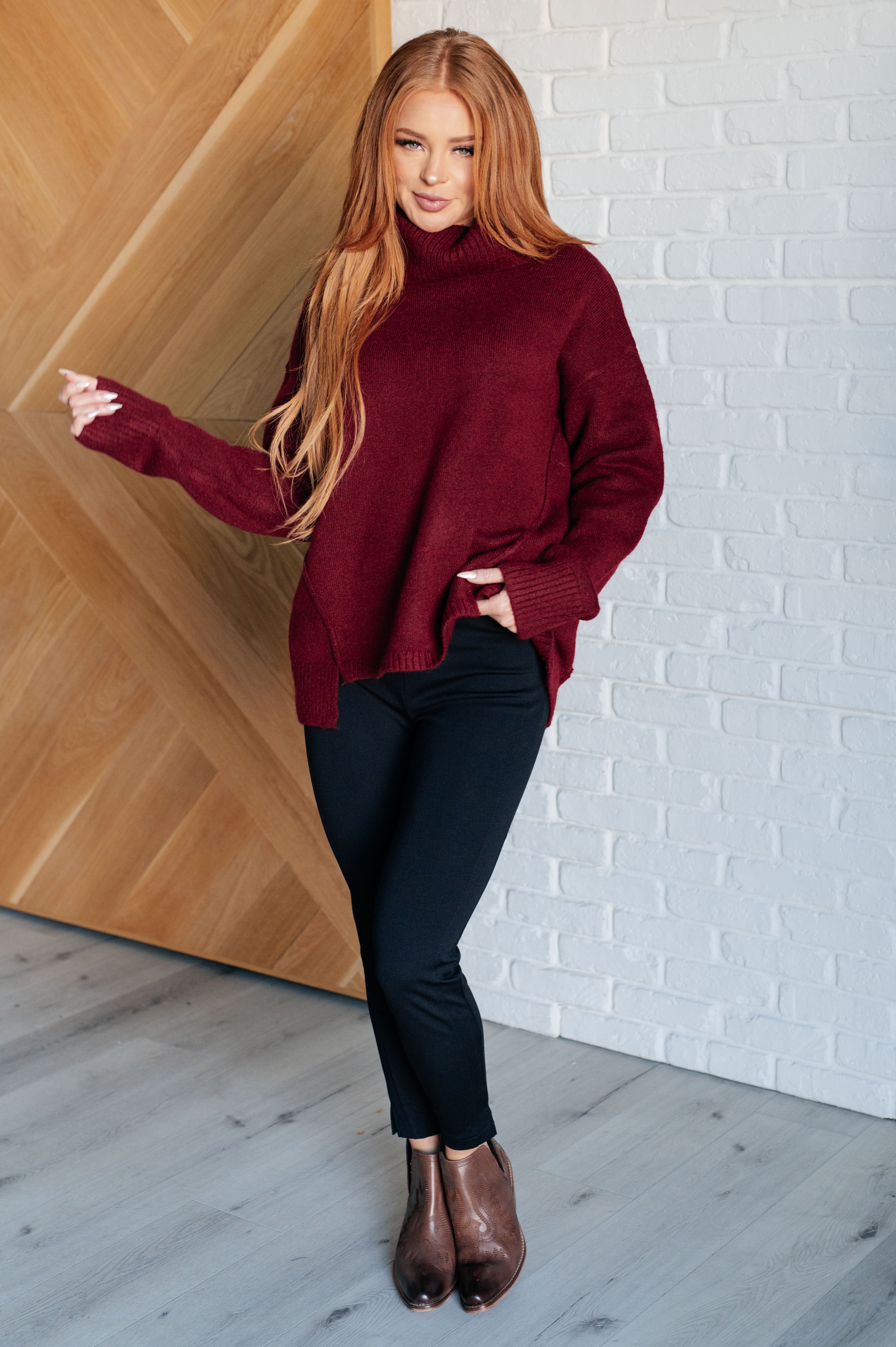 Woman wearing oversized dark burgundy turtleneck sweater with long sleeves paired with black pants and brown boots indoors