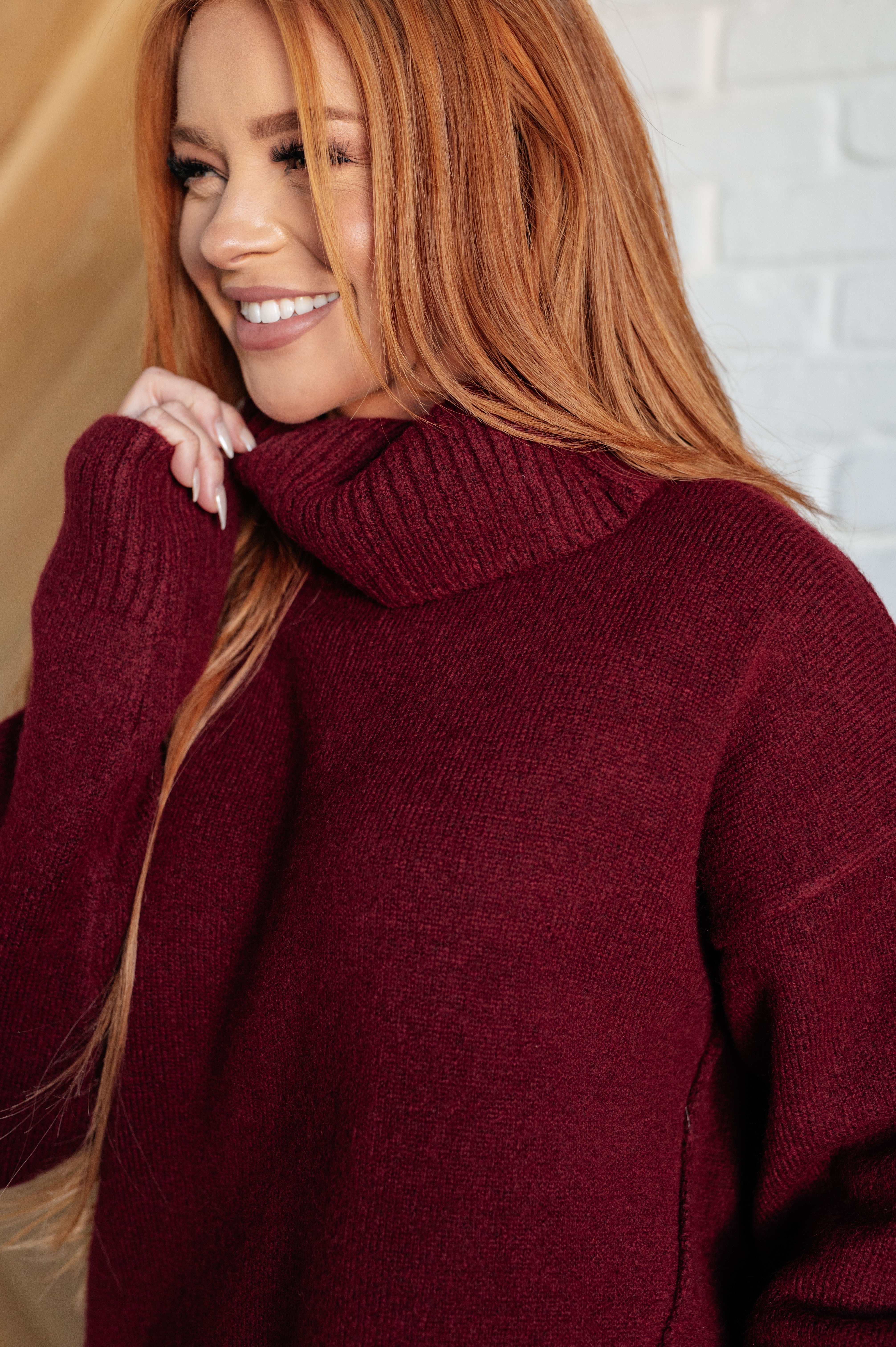Smiling woman wearing deep burgundy turtleneck sweater with long sleeves indoors against neutral background