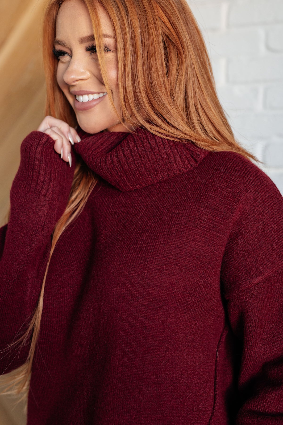 Woman smiling in a burgundy soft knit turtleneck sweater with long sleeves and a relaxed fit indoors beside a white brick wall