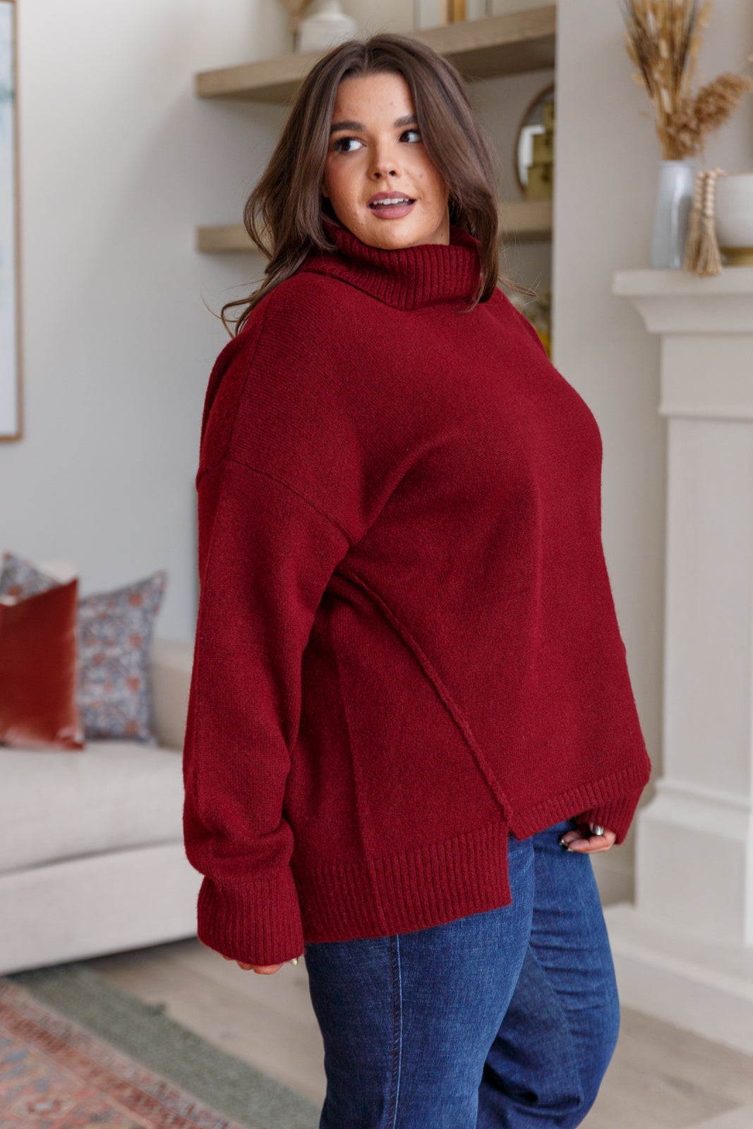 Woman wearing oversized deep red turtleneck sweater paired with blue jeans standing indoors in cozy room