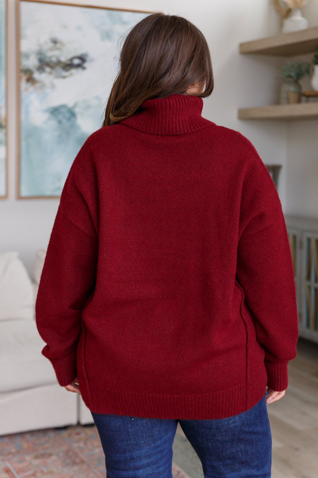 Woman seen from behind wearing deep red turtleneck sweater with long sleeves and blue jeans indoors