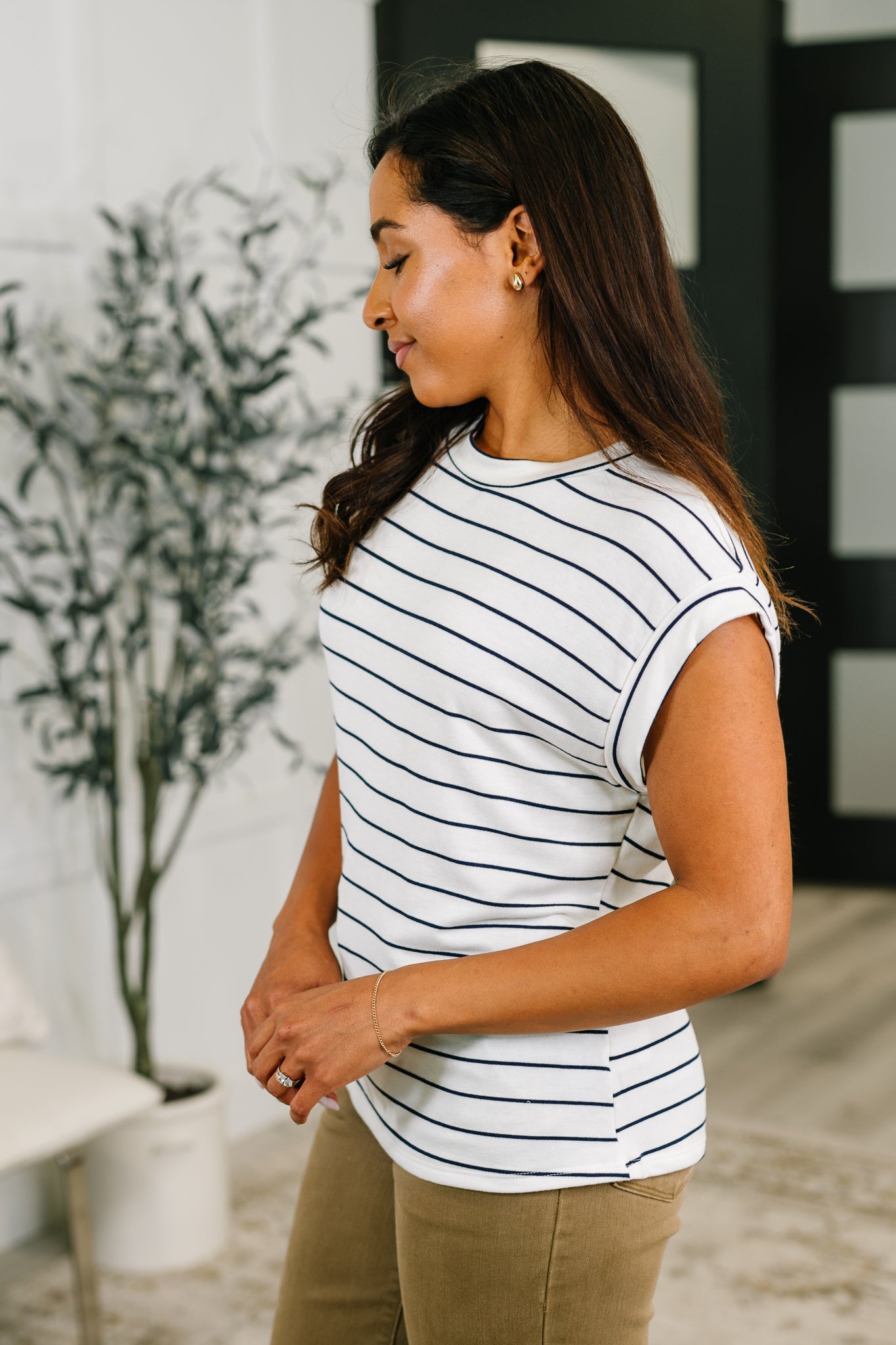Woman wearing an ivory striped cap sleeve tee paired with tan pants standing in a bright eclectic room