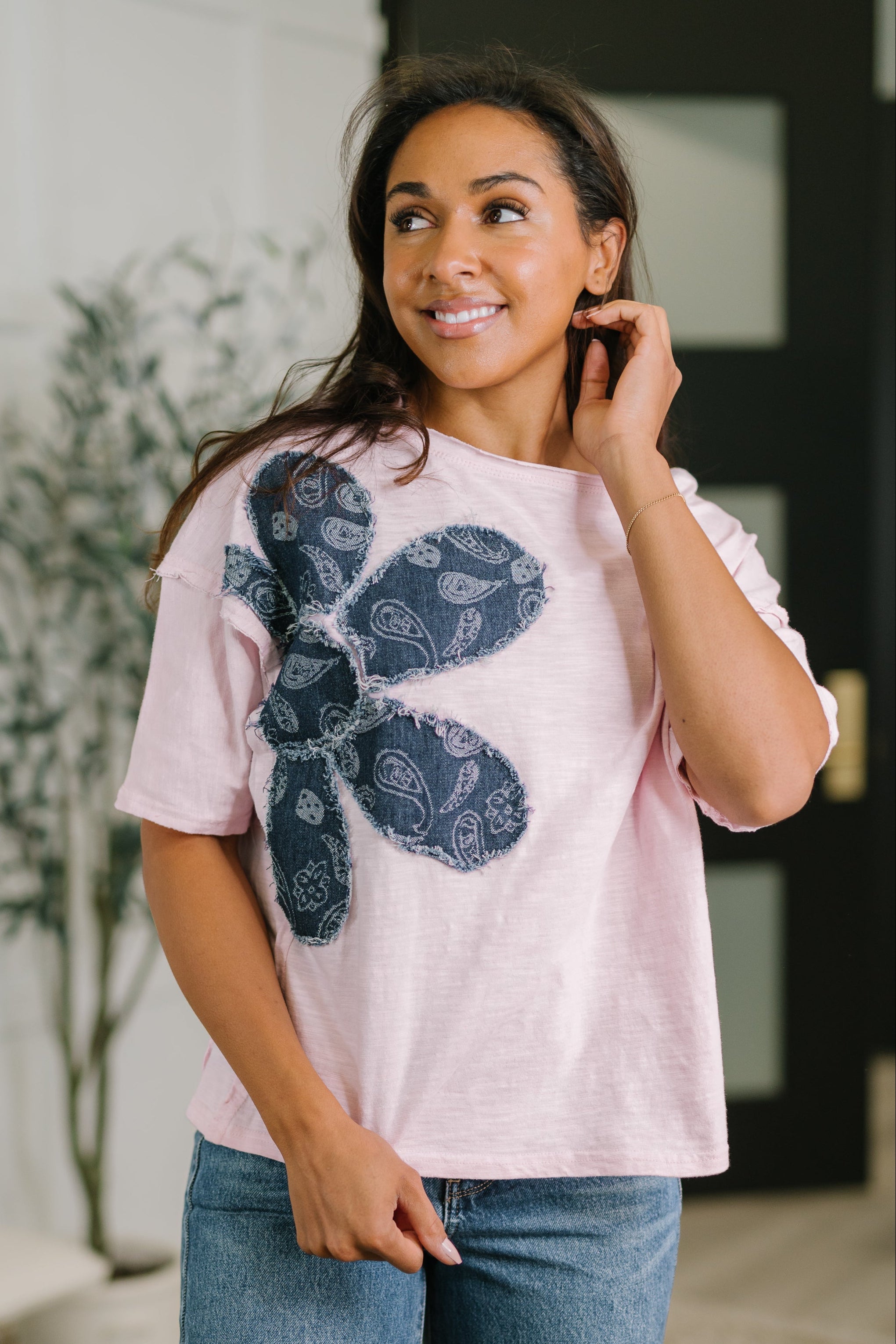 Smiling woman wearing a pink relaxed knit top with dark blue paisley floral applique and casual jeans indoors