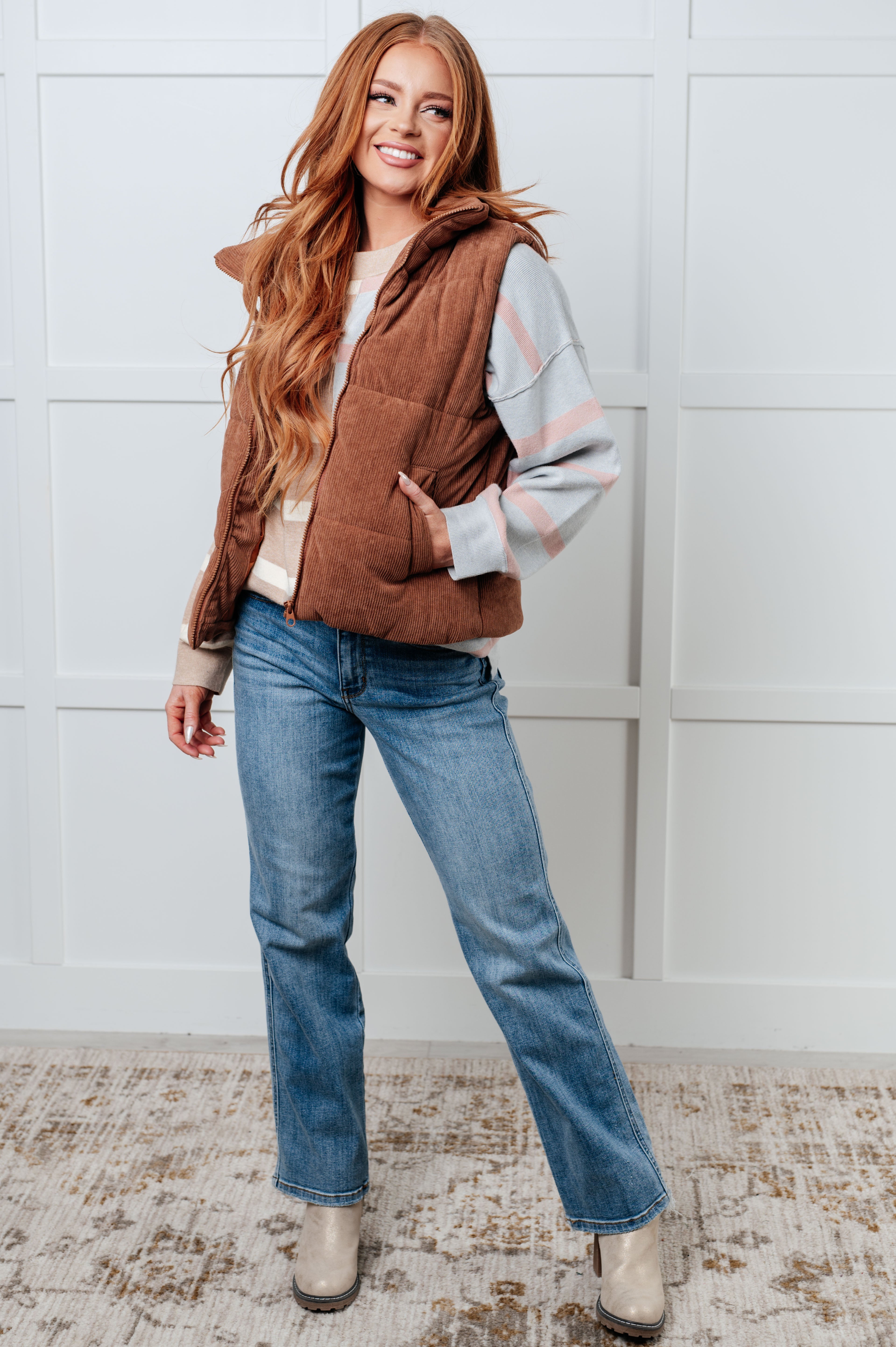 Woman wearing brown quilted vest over striped sweater with light blue jeans and beige boots standing indoors smiling