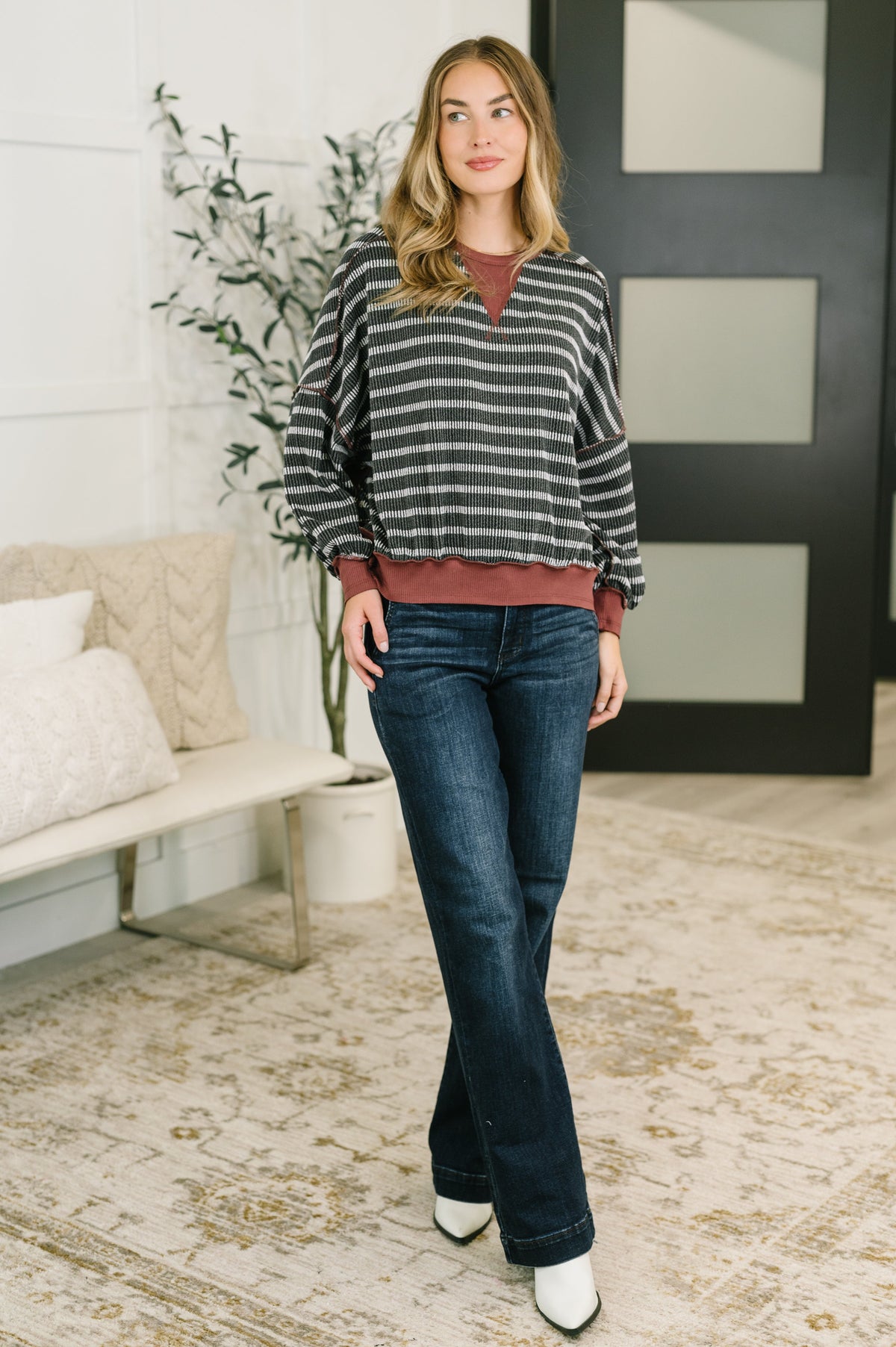 Woman wearing a charcoal contrast textured knit pullover over a rust top with dark jeans and white shoes in a bright home interior