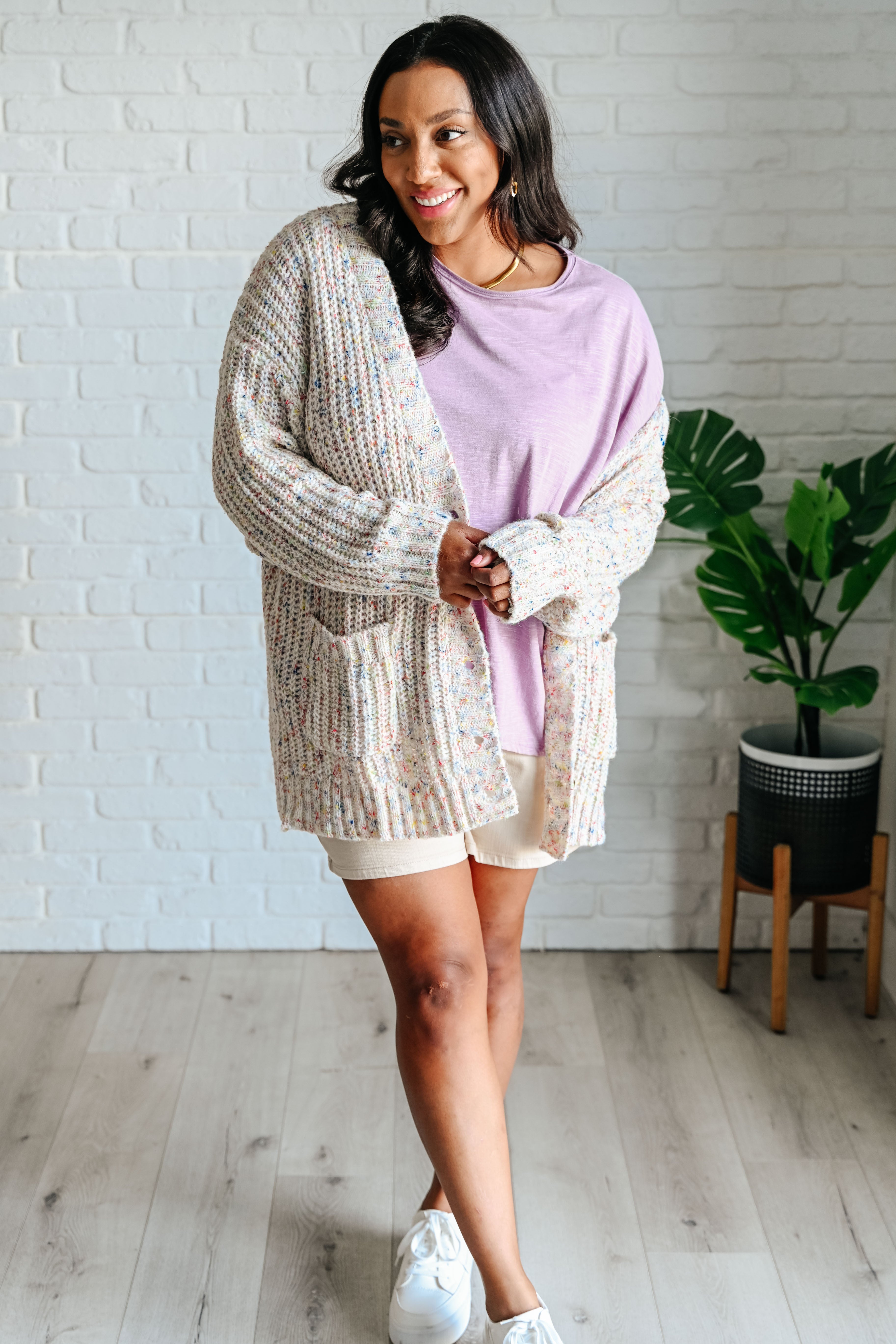 Woman wearing a lavender long sleeve step hem top and a beige confetti cardigan, beige shorts and white sneakers standing in front of a white brick wall