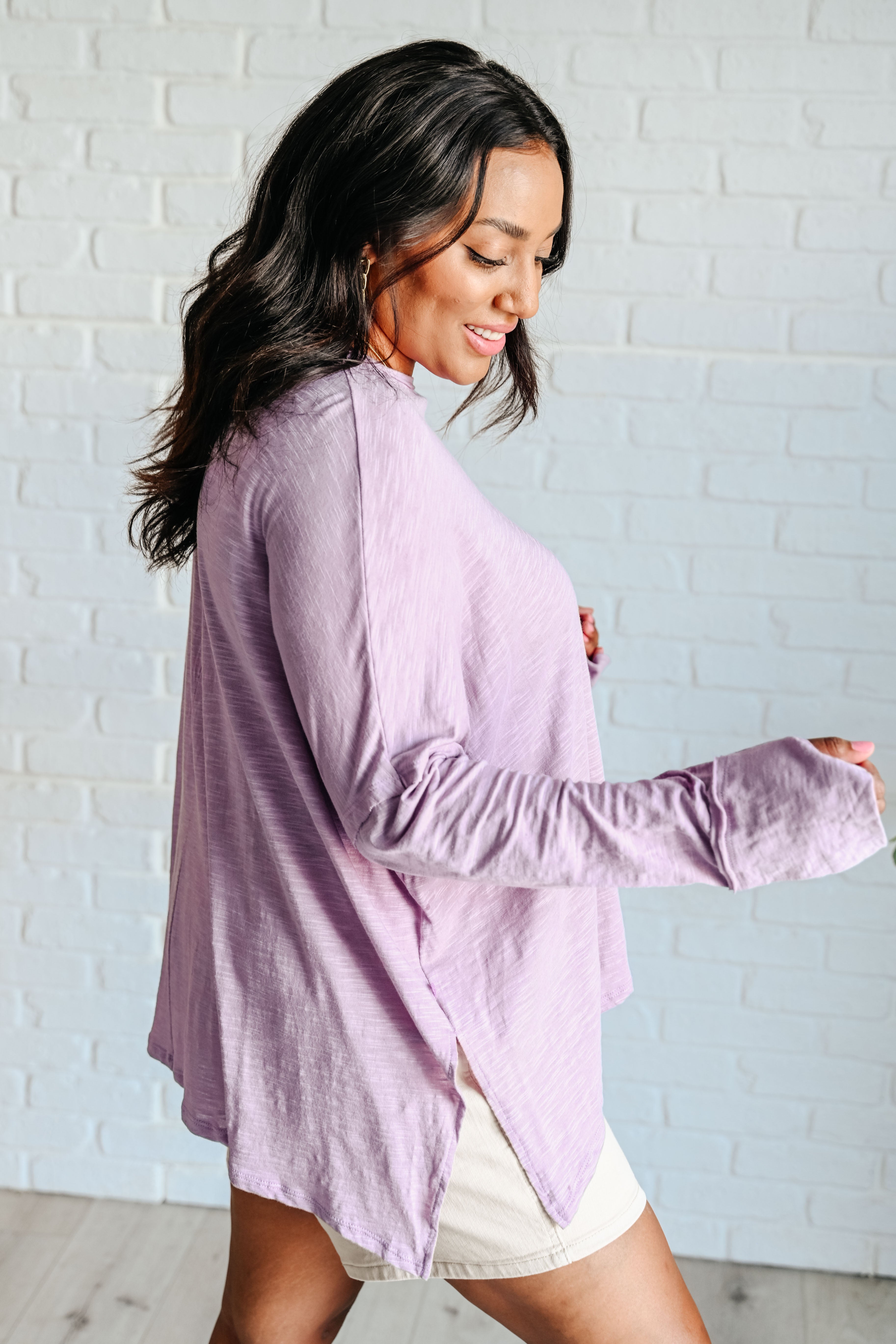 Profile of a woman wearing a lavender long sleeve step hem top over beige shorts standing in front of a white brick wall