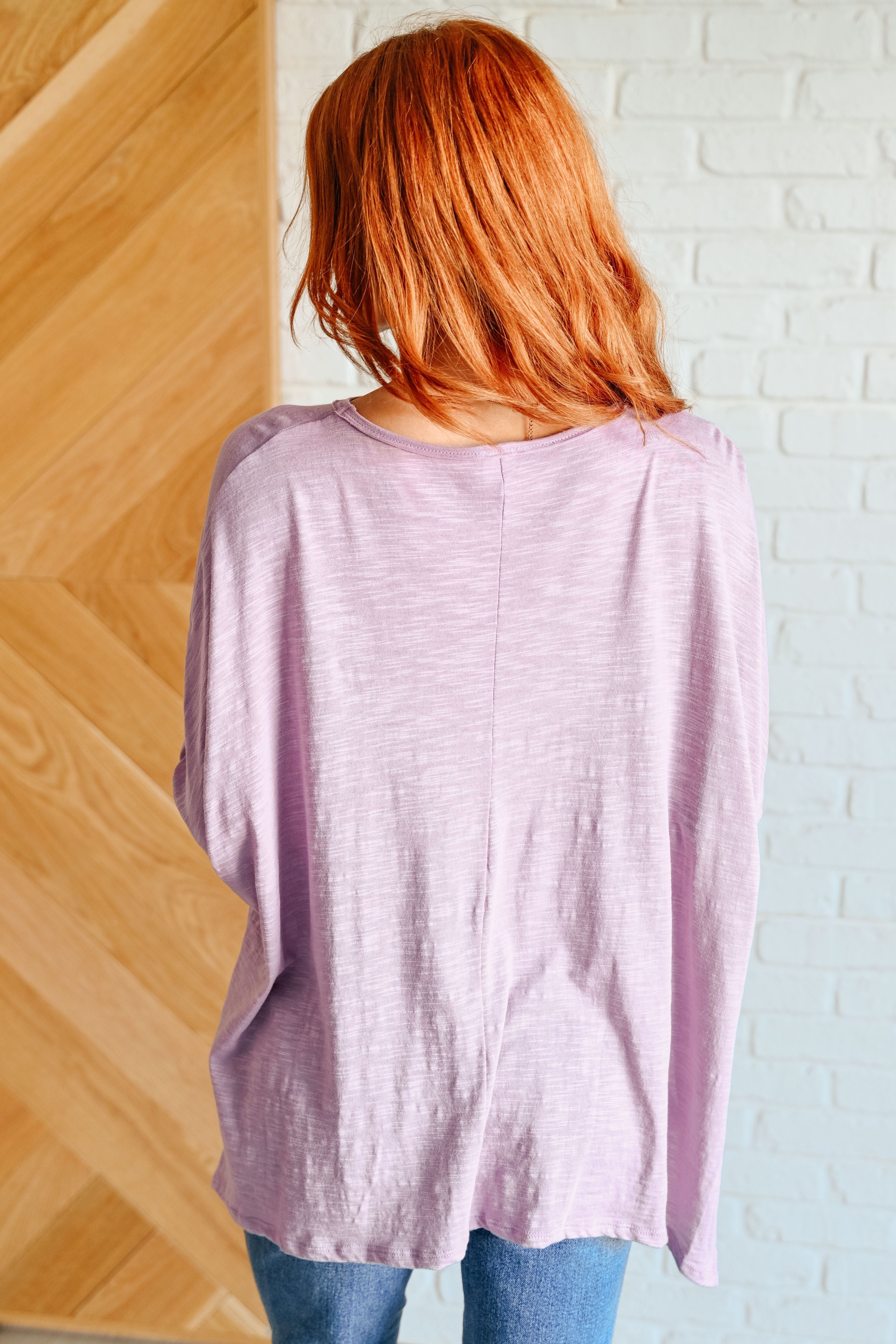 Back facing red haired woman wearing a lavender long sleeve step hem top over blue jeans standing in front of a white brick wall