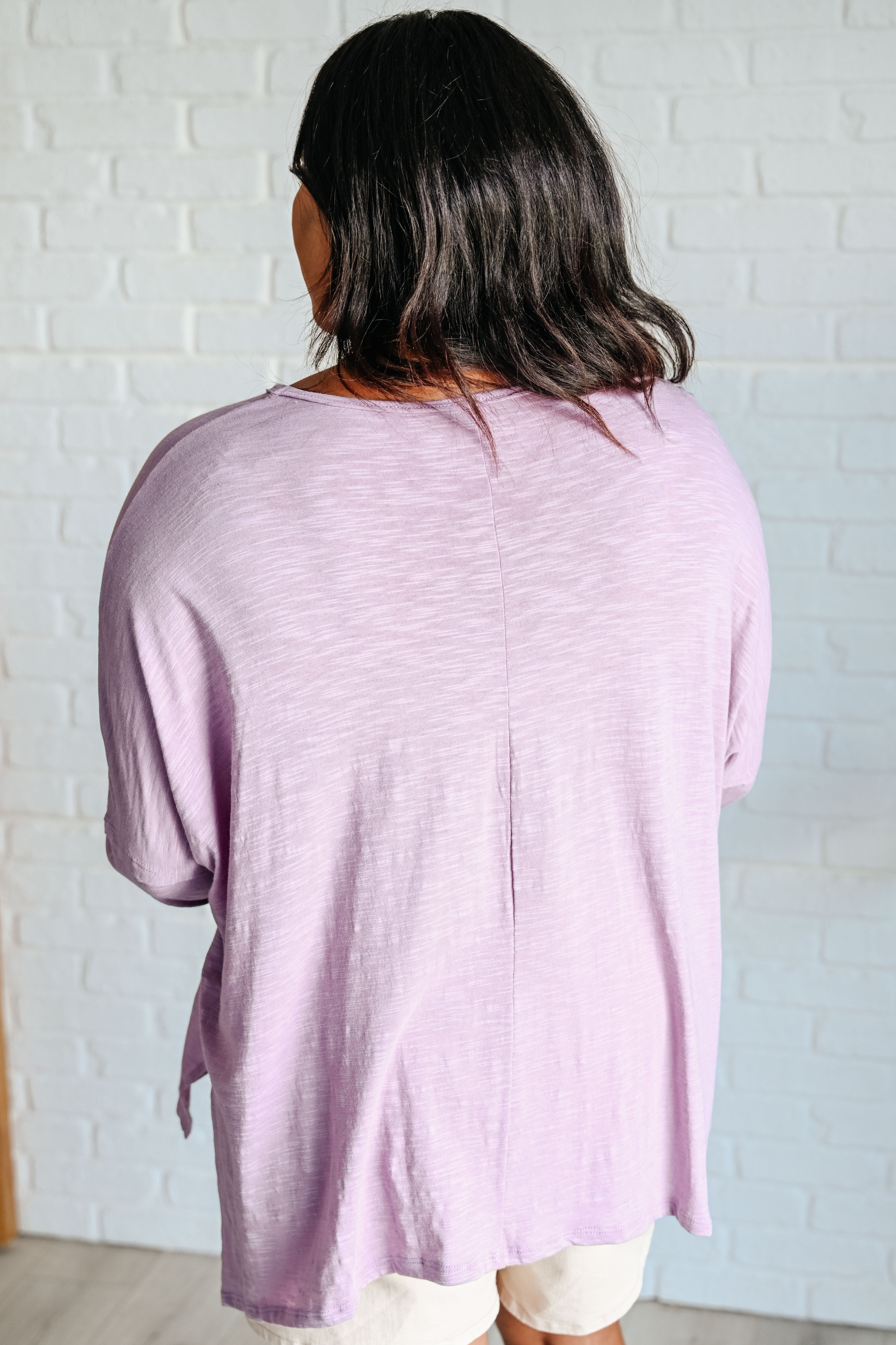 Back facing woman wearing a lavender long sleeve step hem top over beige shorts standing in front of a white brick wall