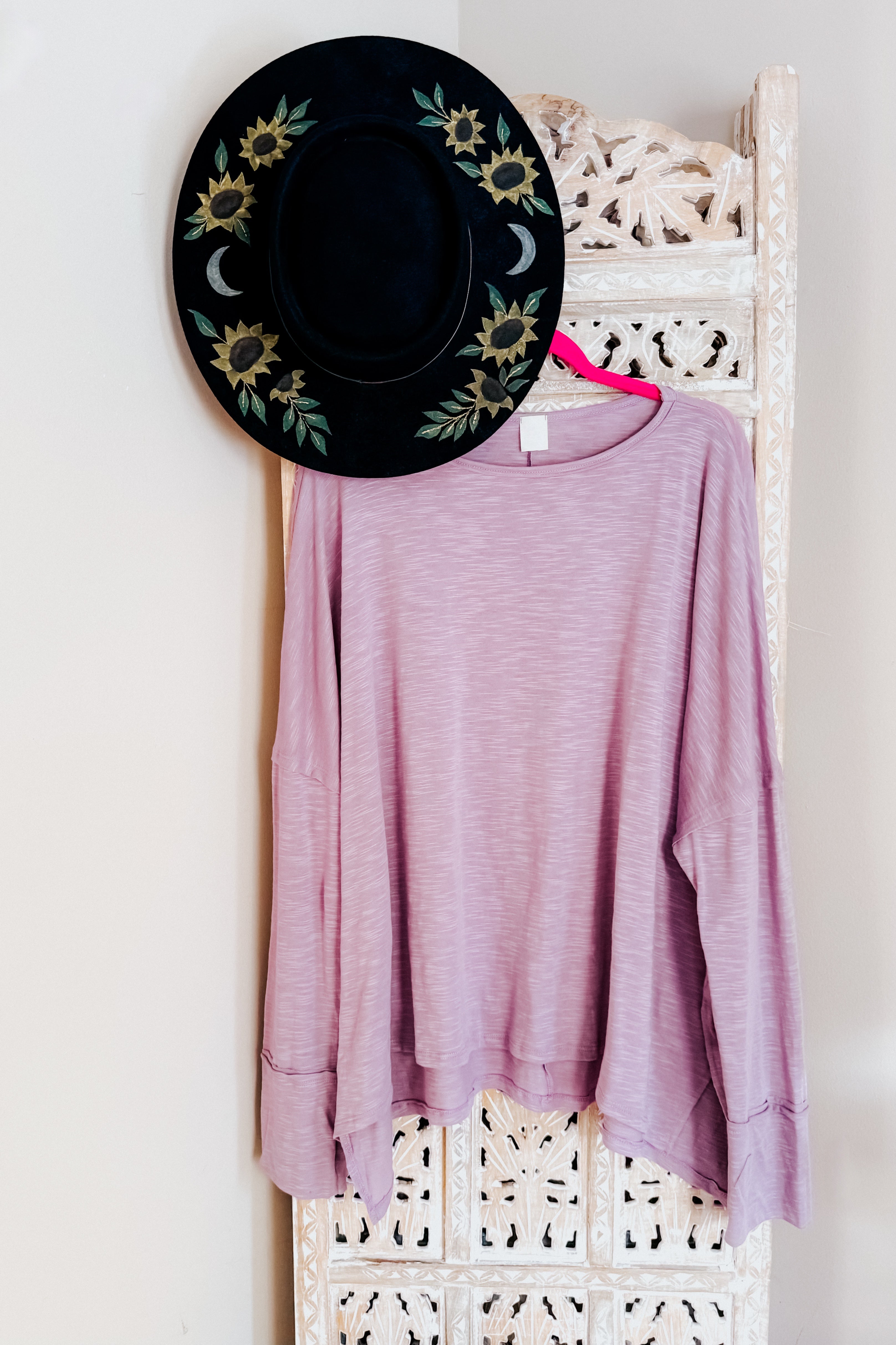 A lavender long sleeve top with a step hem hanging on a ornate room divider with a hand painted wide brim hat
