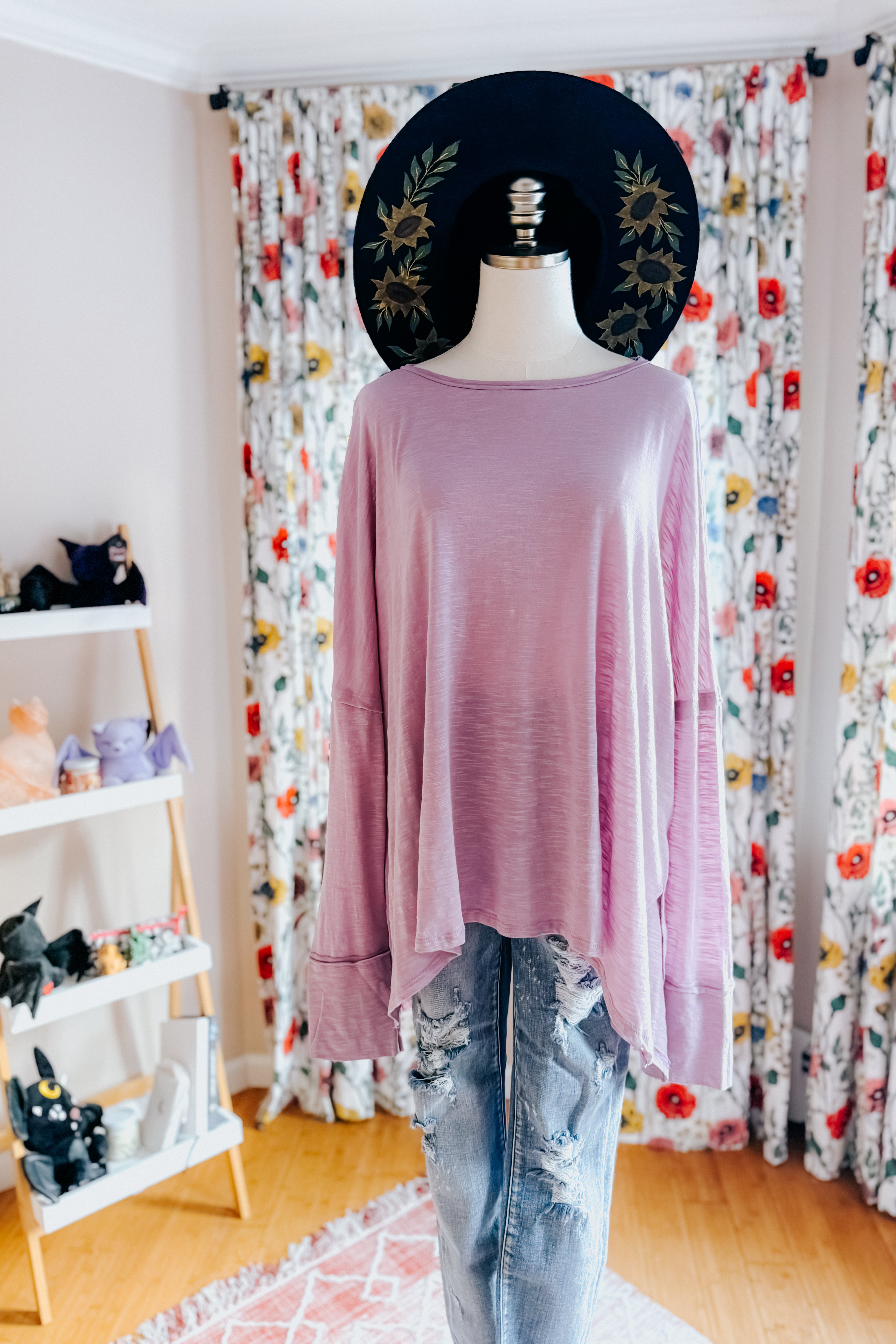 Dress form wearing a lavender long sleeve step hem top, distressed blue jeans and a hand painted wide brim hat in an indoor setting with floral curtains