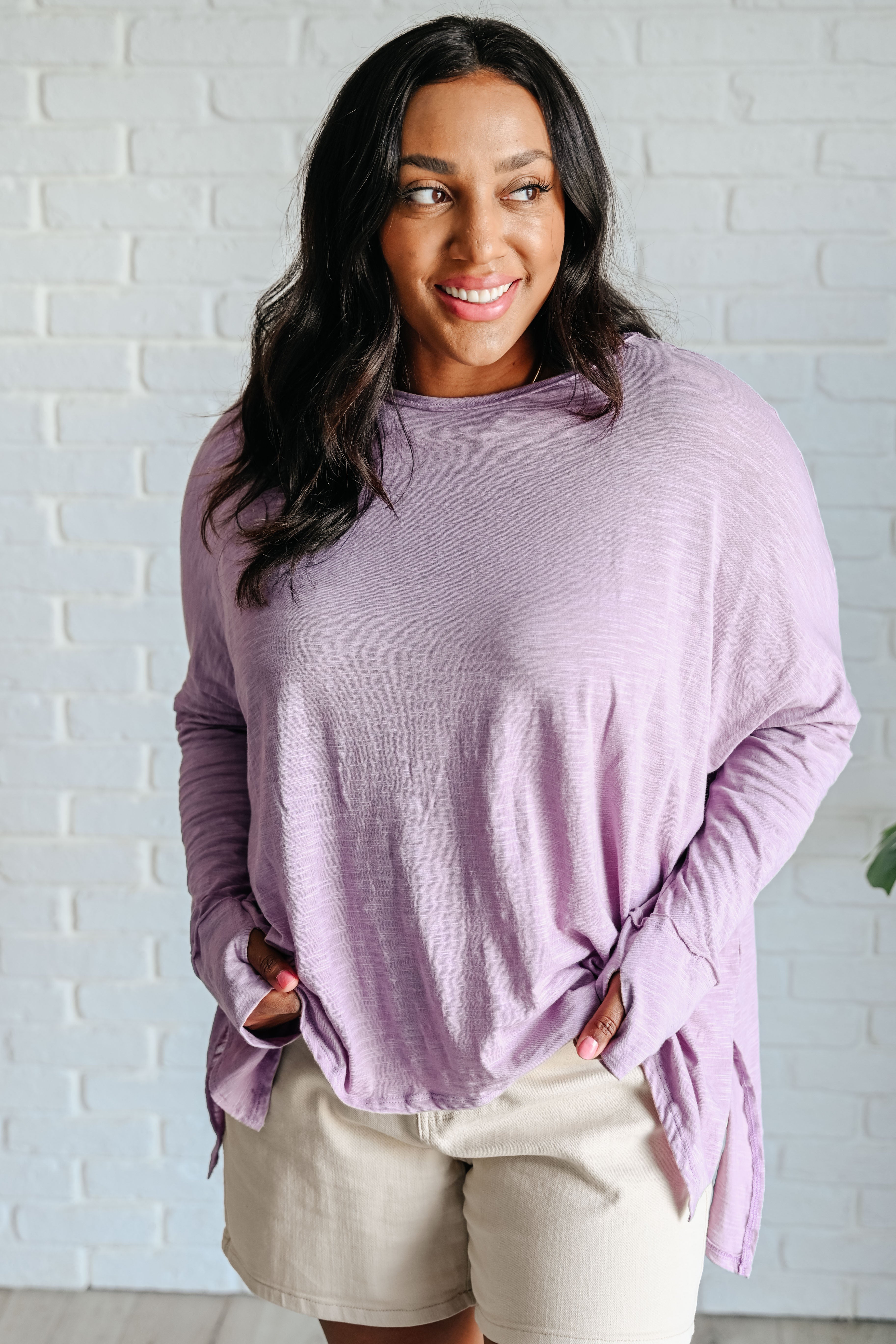 Woman wearing a lavender long sleeve step hem top over beige shorts in front of a white brick wall