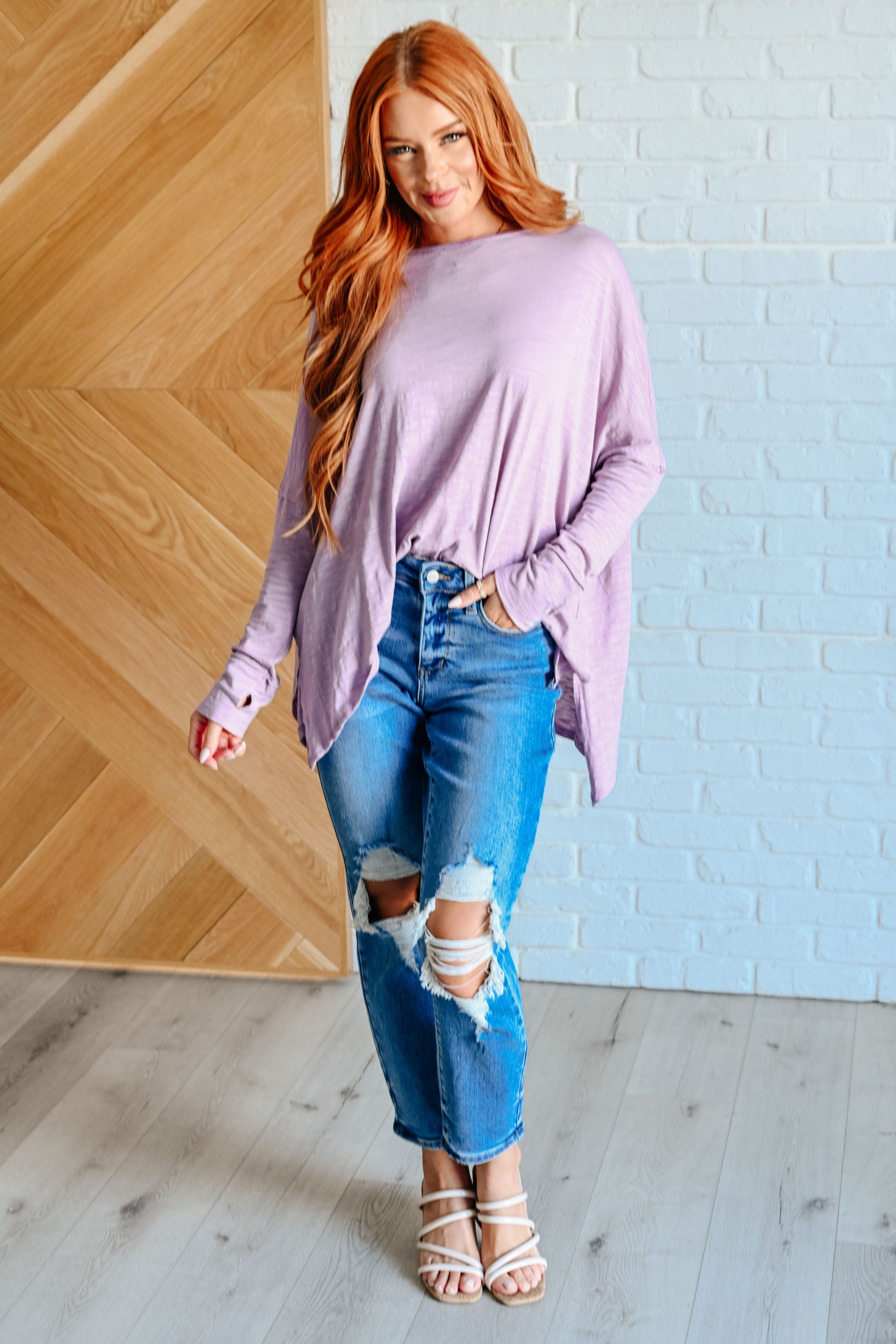 Red haired woman wearing a lavender long sleeve step hem top over blue distressed jeans and white sandals standing in front of a white brick wall