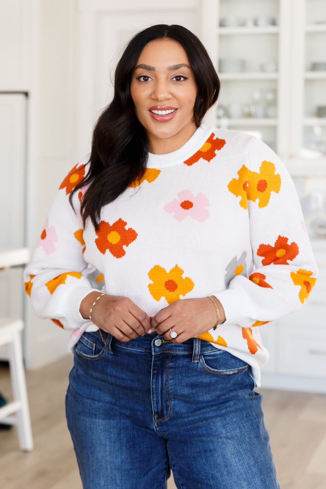 Smiling woman wearing white sweater with orange, yellow, and pink floral designs paired with blue jeans indoors