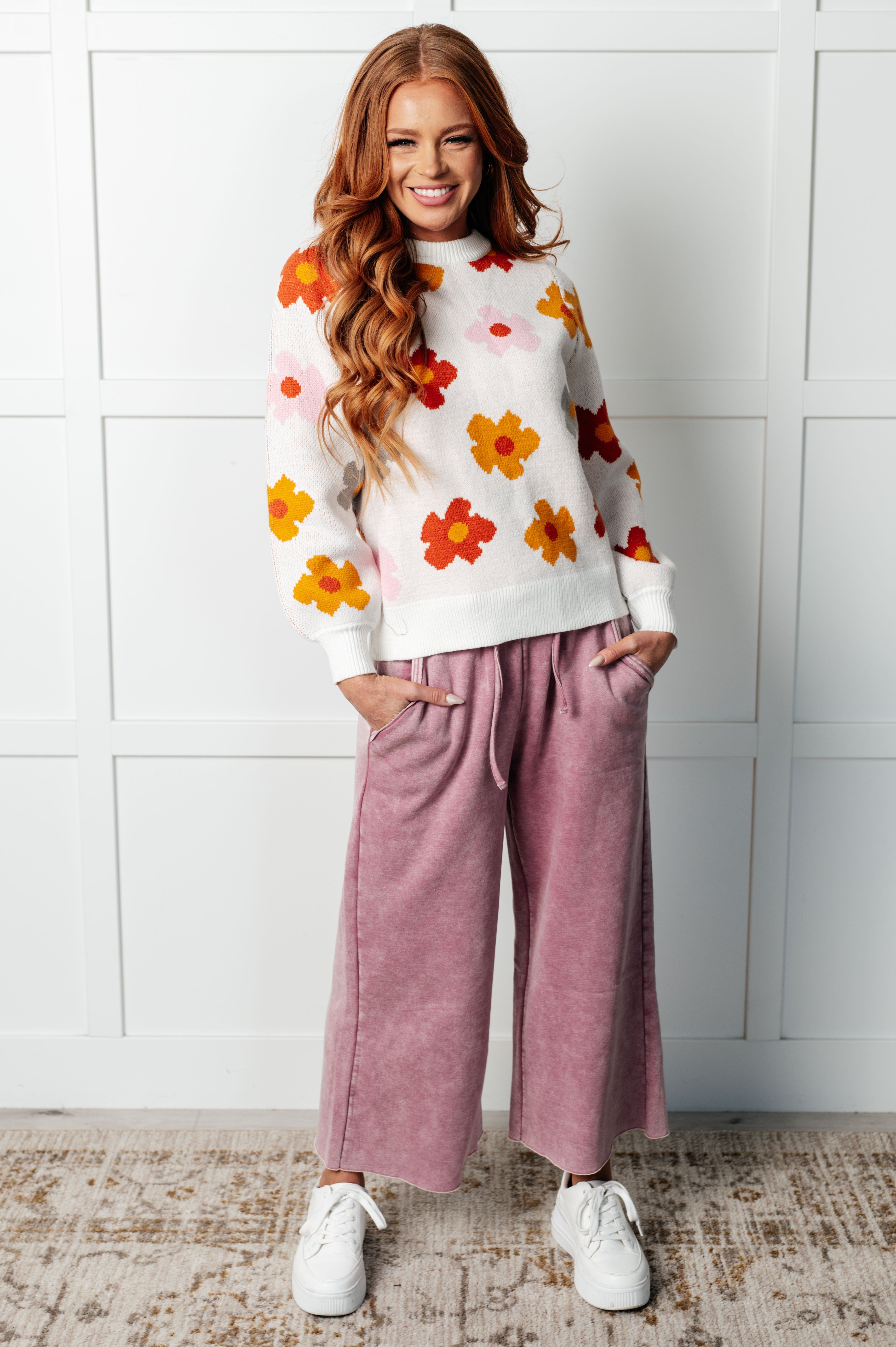 Smiling woman with long red hair wearing white floral sweater with orange and pink flowers paired with mauve wide-leg pants indoors