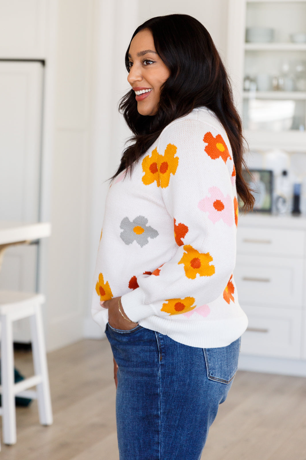 Smiling woman in white sweater with orange, yellow, gray, and pink floral designs paired with blue jeans indoors