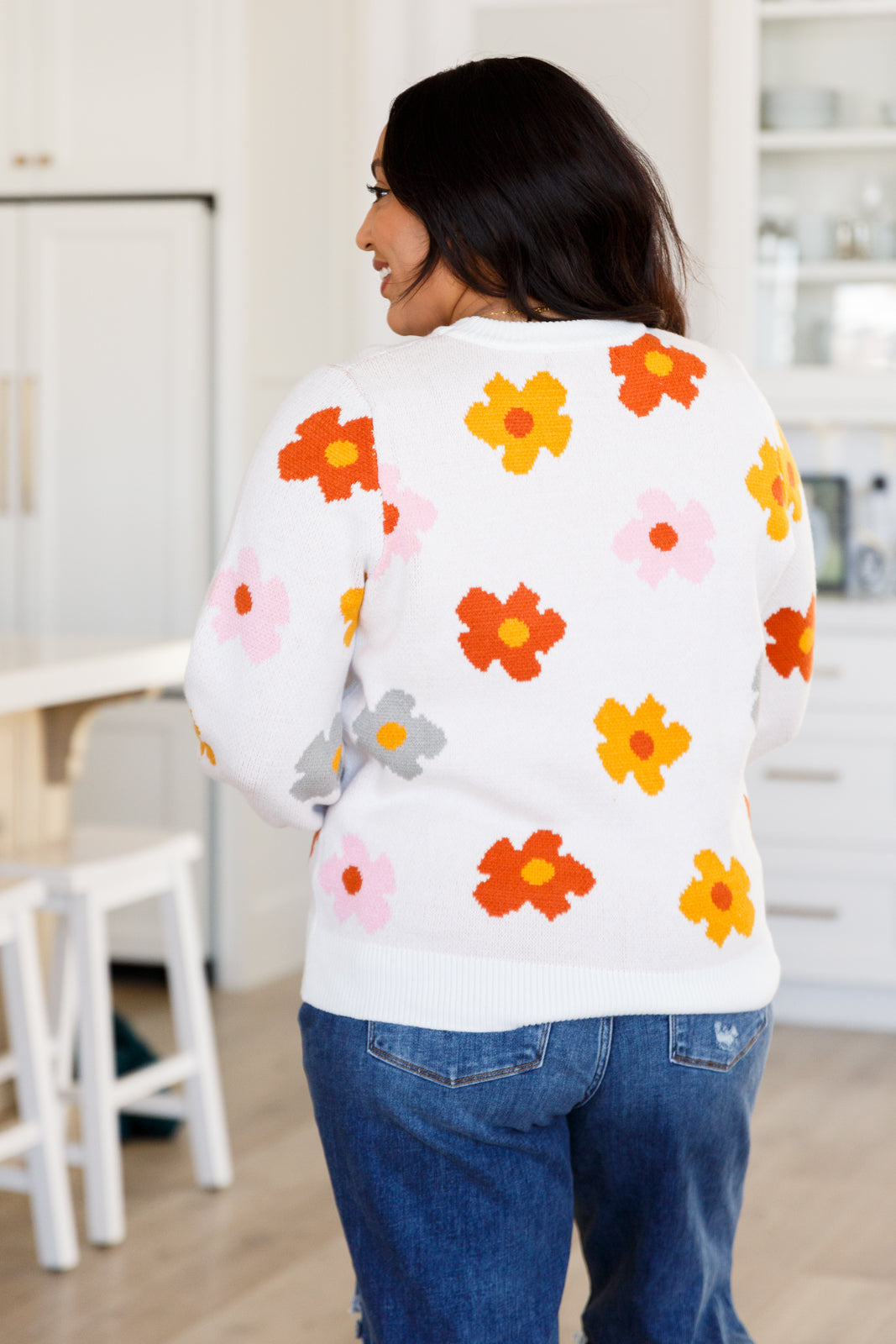Woman wearing white sweater with orange, yellow, pink, and gray floral designs paired with blue jeans indoors from the back