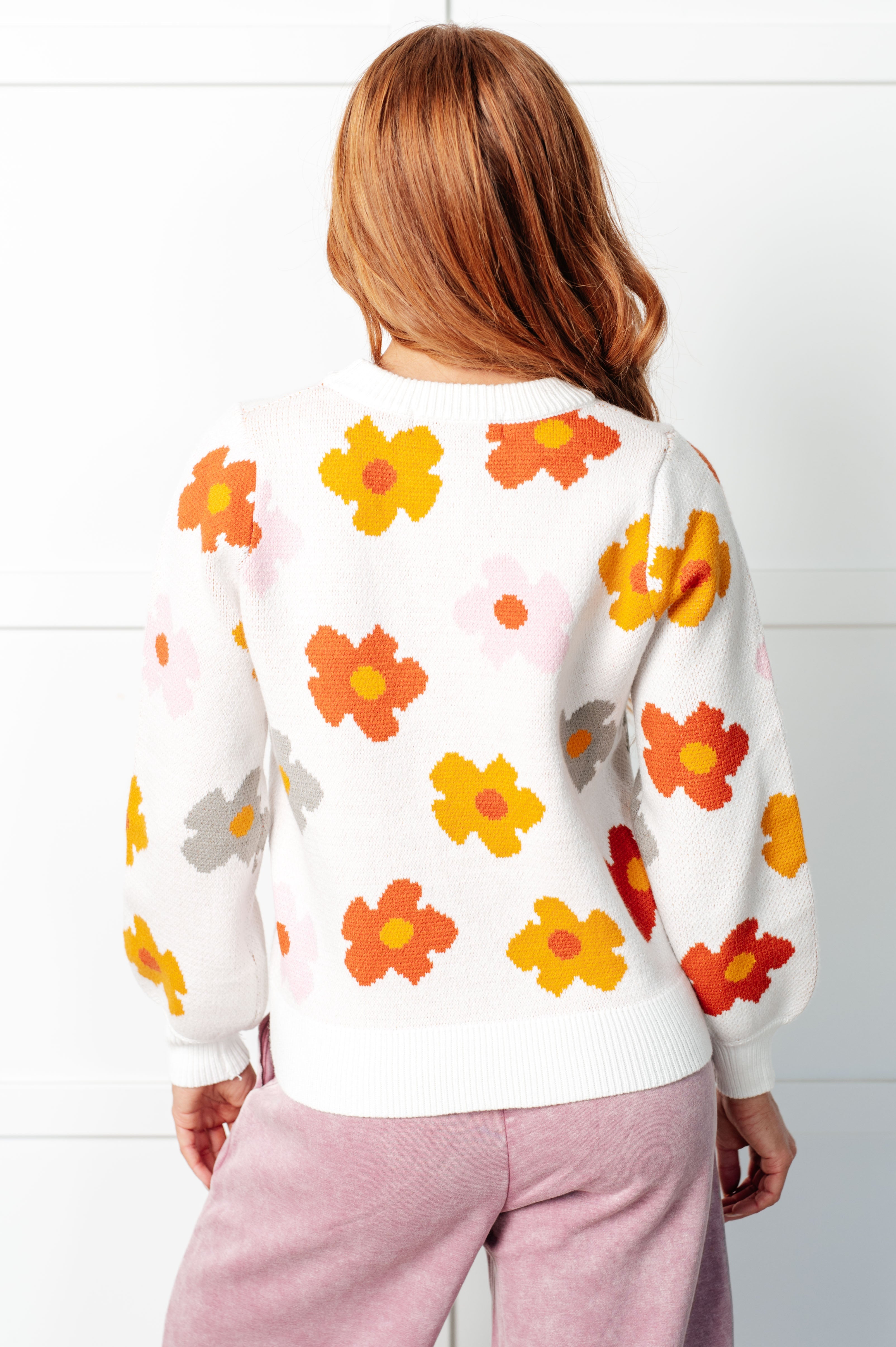 Woman wearing white sweater with bold orange, yellow, pink, and gray floral prints paired with light purple pants indoors from the back