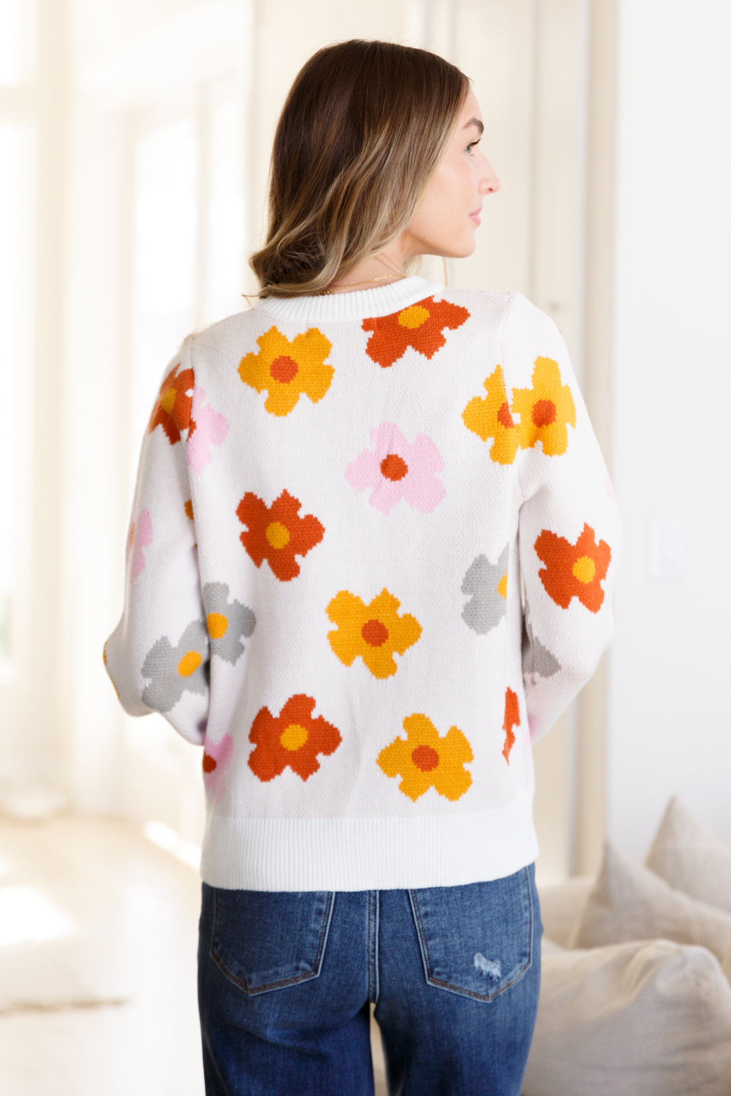 Woman wearing white sweater with bright orange, red, yellow, pink, and gray floral prints paired with blue jeans indoors from the back