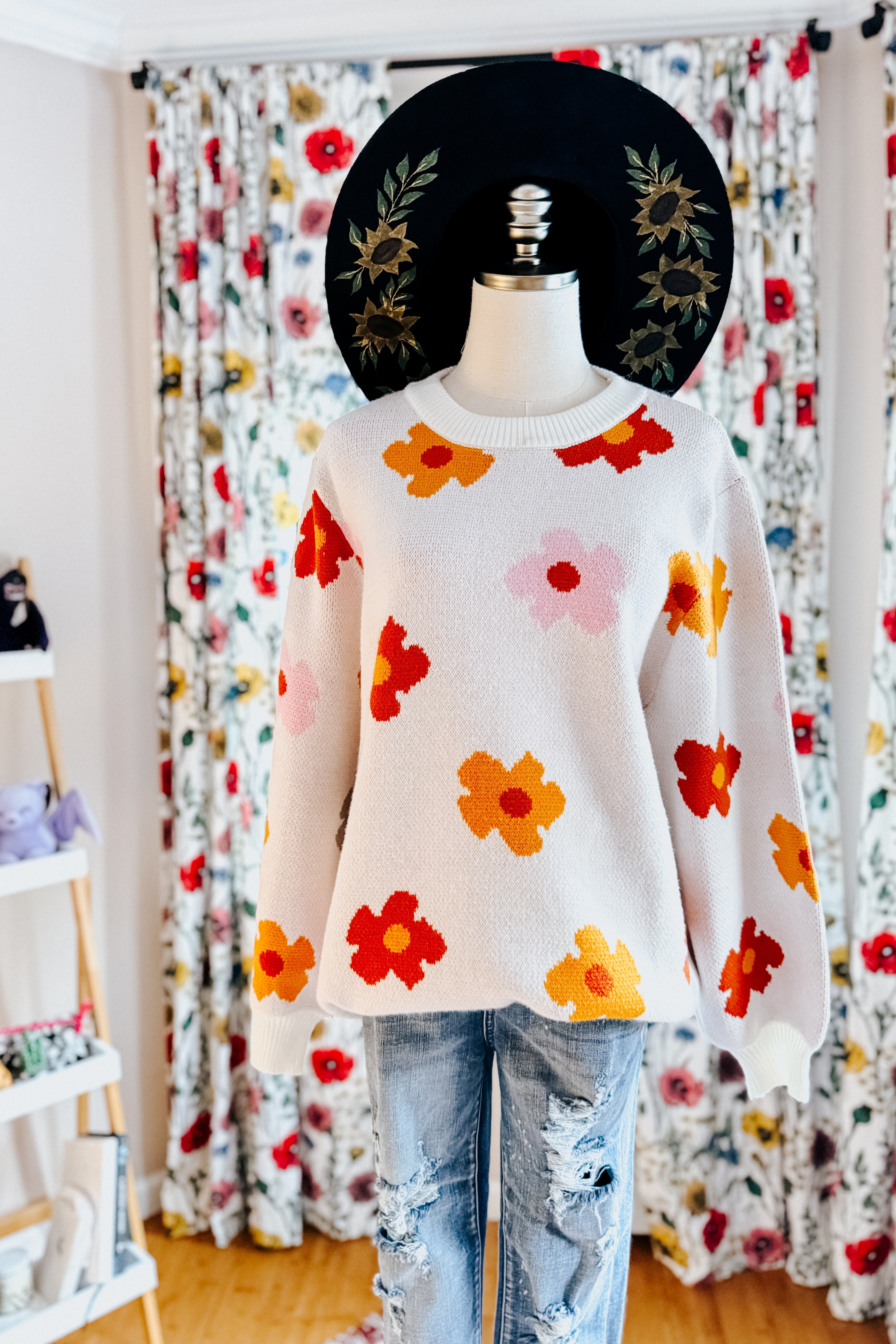 White sweater with bold red, orange, and pink floral patterns paired with ripped light blue jeans on mannequin indoors