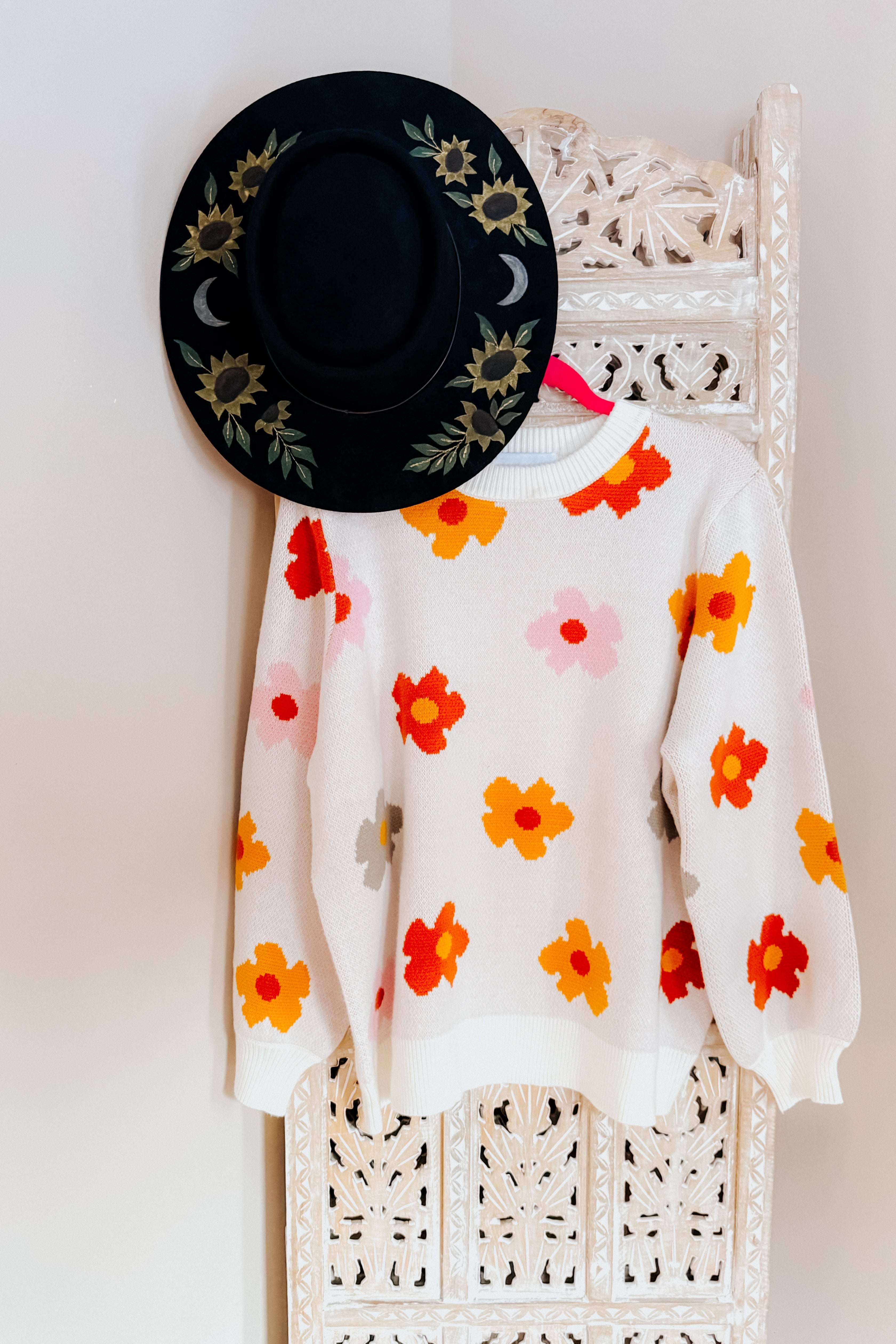 Cream sweater with bright orange, red, and yellow floral print hanging on carved white wooden panel with black floral hat above