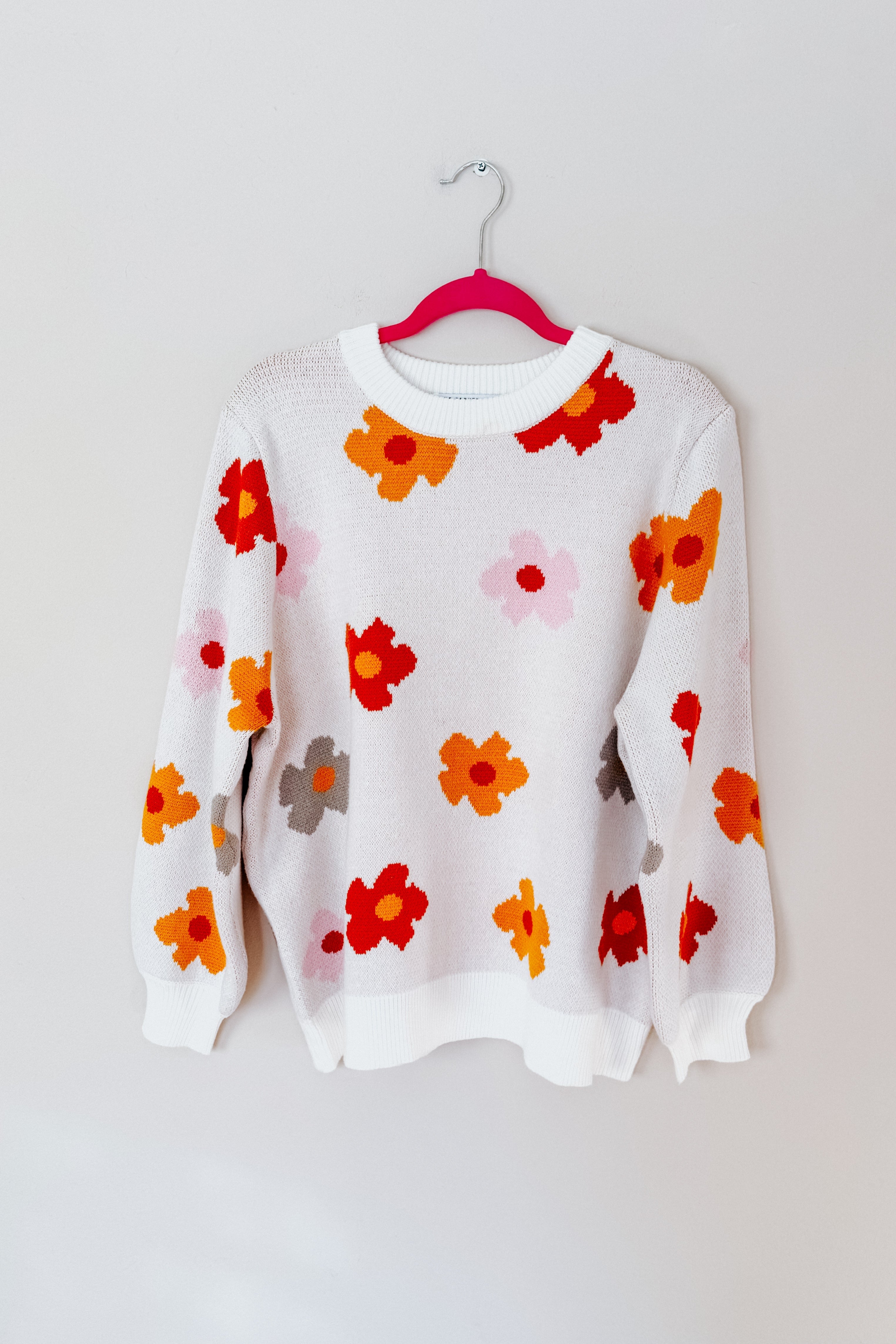 White knit sweater with scattered large floral patterns in red, orange, pink, and gray hanging on pink hanger against white wall